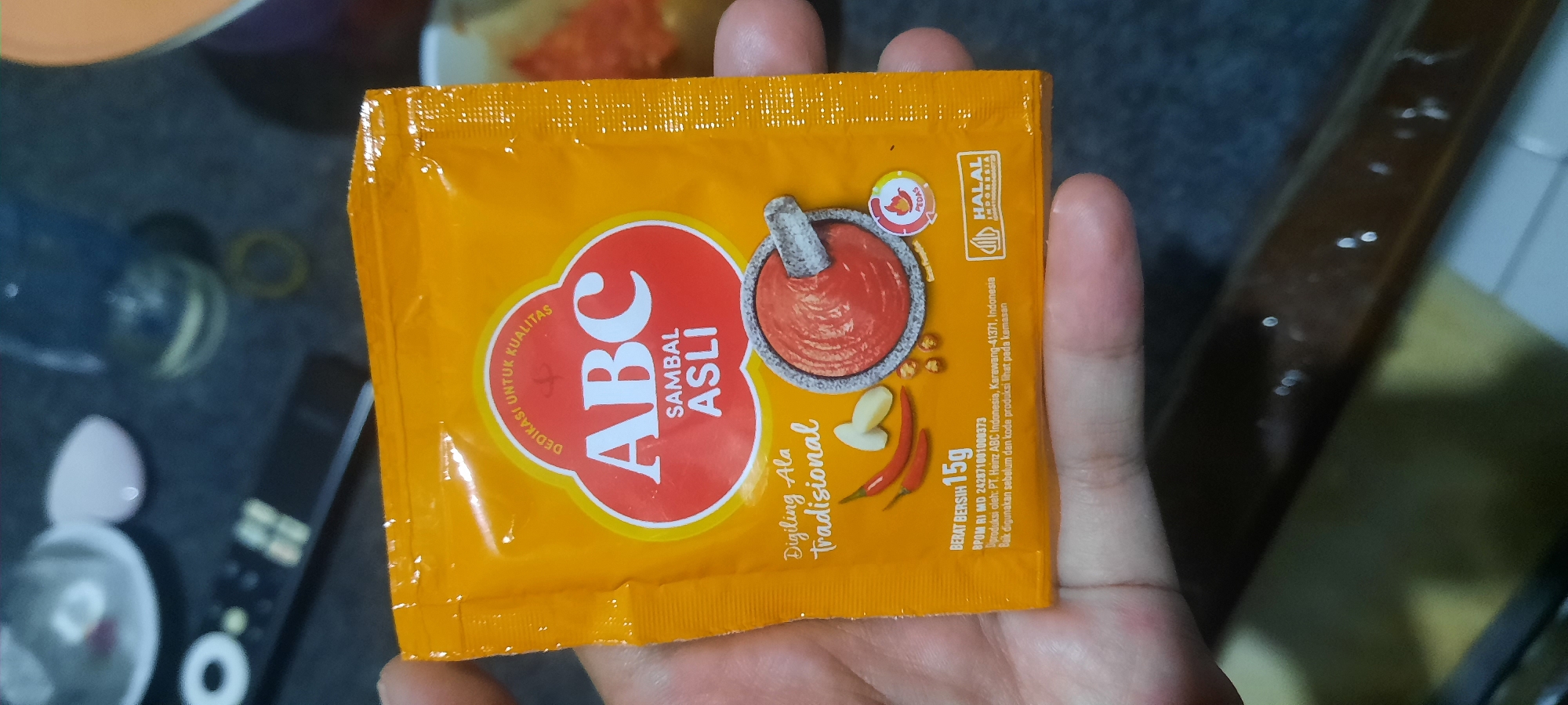 An image shows a hand holding a packet of ABC Sambal Asli, a brand of chili paste from Indonesia. The packet is orange and features the ABC logo, along with text indicating "Sambal Asli" (Original Chili Paste). Below the logo, there is an illustration of chili peppers and garlic being pounded in a mortar and pestle, representing a traditional preparation method. The packet also displays "Berat Bersih 15g" (Net Weight 15g), BPOM RI MD number, and production details indicating it is produced by PT. Heinz ABC Indonesia in Karawang, Indonesia. A Halal logo is also visible on the packet. The background appears to be a dark, somewhat blurry surface with some objects that are not clearly identifiable. The image does not provide any specific details or context related to the city of North Jakarta.