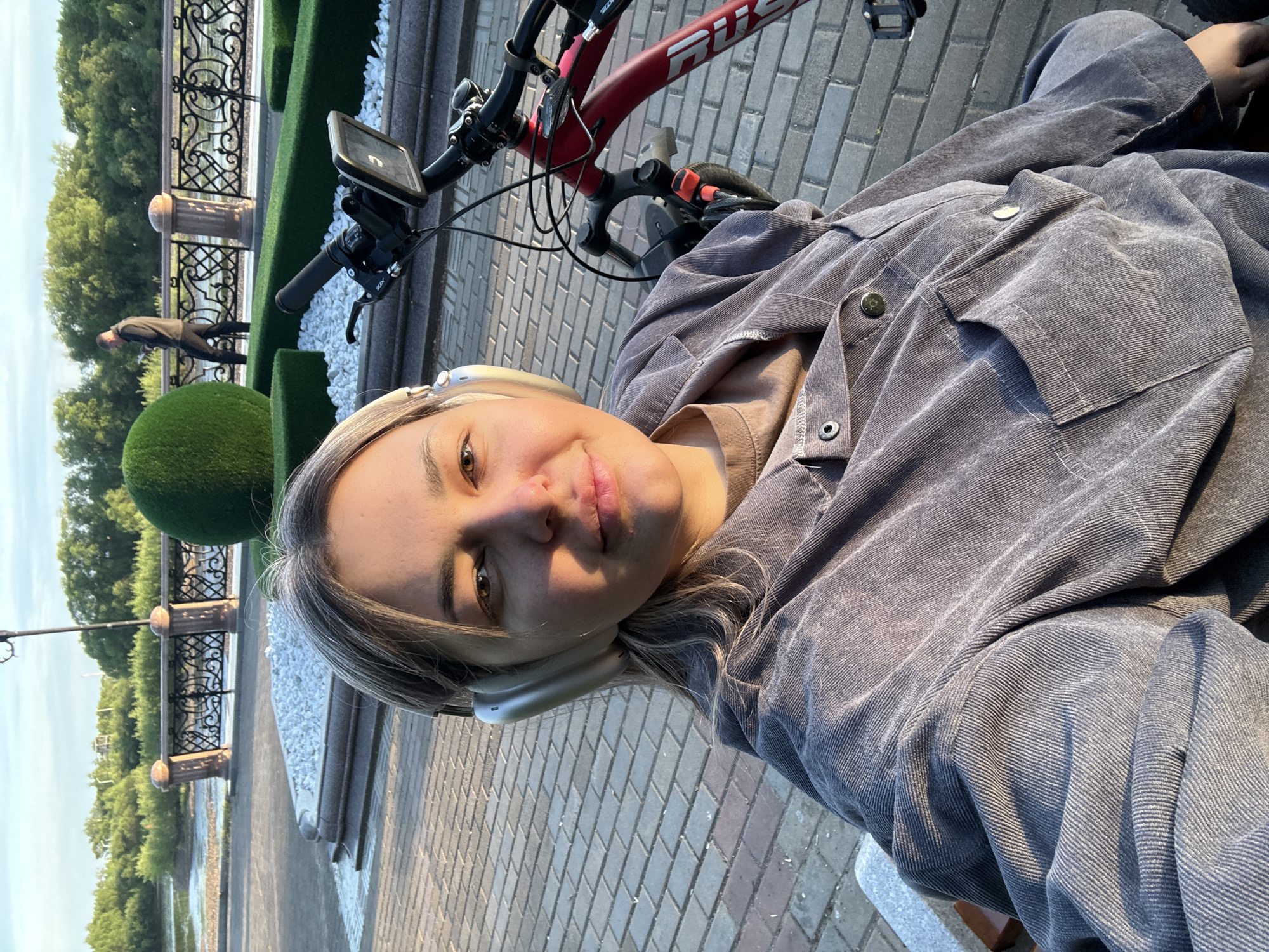 A waist-up selfie shows a woman with short blonde hair, wearing grey headphones and a grey corduroy button-up shirt, with a red bicycle partially visible above her. The background shows an outdoor urban scene. There is a patterned iron railing with concrete posts and a trimmed hedge. Behind the hedge, a person is walking and there is a manicured topiary ball. The area looks like a park or a promenade, with a body of water visible beyond the railing. The pavement is made of grey brick tiles. The woman is likely sitting, as the angle is looking down on her. It is impossible to determine whether the location is the city of Birobidzhan, Russia.