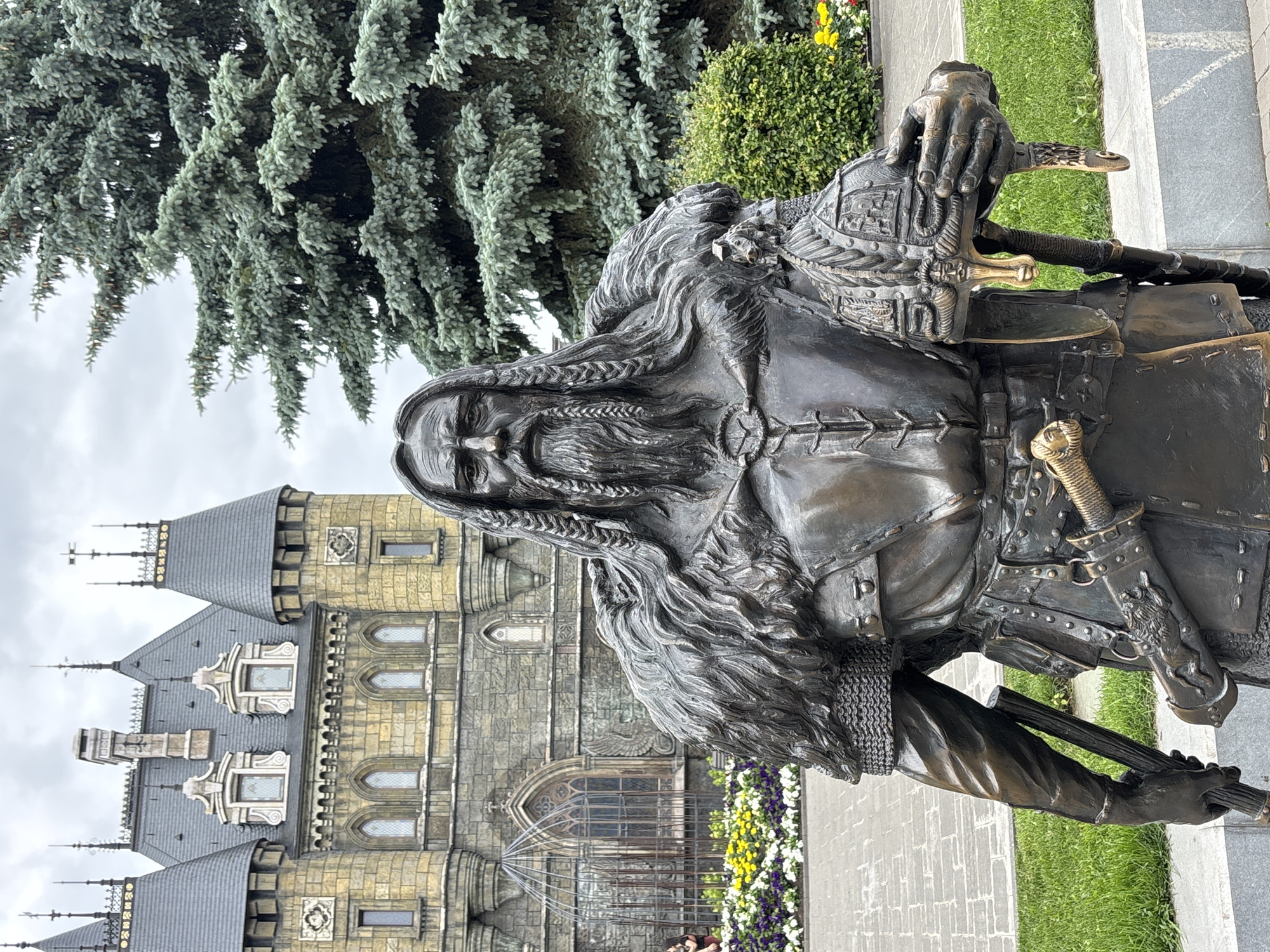 The image features a bronze statue of a historical figure, likely a warrior or leader, positioned in front of a castle-like building. The statue depicts a man with a long beard and braided hair, wearing armor and a fur cloak. He is holding a helmet in his right hand and a sword is sheathed at his hip. In the background, a large, ornate building with turrets and arched windows is visible. To the right of the statue, a large pine tree stands tall. The foreground shows a paved walkway, a grassy area, and a flowerbed with colorful flowers. The sky is overcast, suggesting a cloudy day. This scene is located in Khryashchevka, Russia, at the "Fairy Tale Forest" park, which is known for its replicas of medieval structures and statues.