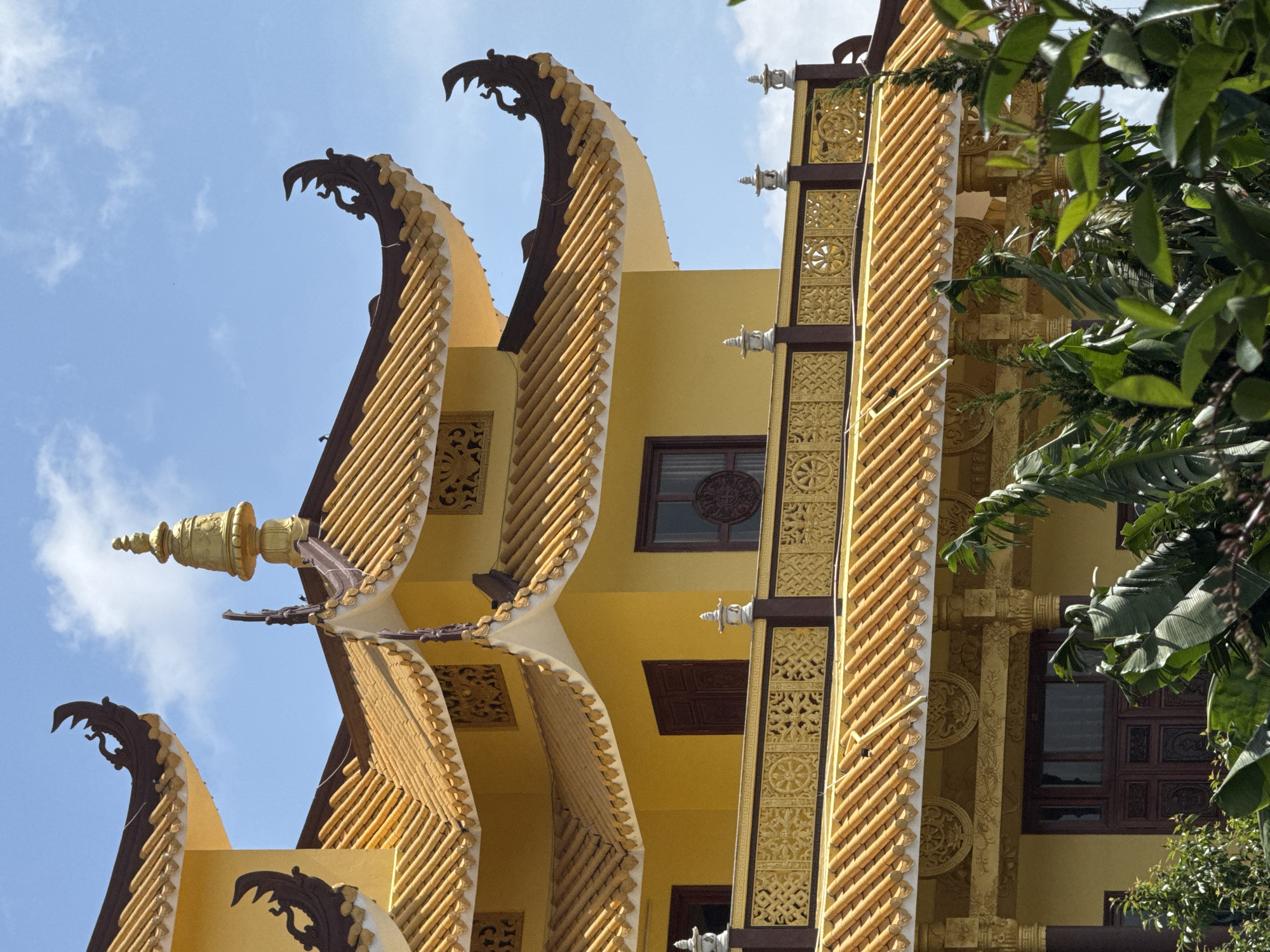 This image depicts a vibrant yellow Buddhist temple, likely the **Chua Thien An temple in Da Lat, Vietnam**, with its distinctive tiered roofs and ornate decorations. The traditional East Asian architectural style is evident in the curved eaves, topped with dark, decorative elements that resemble stylized dragons or mythical creatures. Golden tiles cover the roofs, catching the sunlight. The building itself is painted a bright yellow, a color often associated with royalty and spirituality in Asian cultures.
A prominent feature is the golden stupa-like finial crowning the highest roof section. Below this, a balustrade with intricate golden carvings runs along a balcony, adorned with decorative lamps or finials. The carvings on the balustrade feature geometric patterns and what appear to be Dharma wheels, a significant Buddhist symbol. Dark brown wooden windows are visible on the building's facade. Lush green foliage, including large-leafed plants and some evergreen trees, partially obscures the lower sections of the temple, adding a natural element to the scene. The sky above is a clear blue, punctuated by scattered white clouds, suggesting a bright, sunny day. No people are visible in the frame.