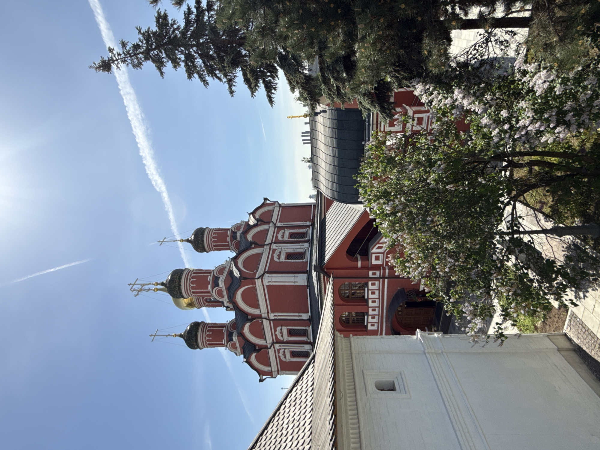 The image captures a view of a red and white Russian Orthodox church against a blue sky with a visible contrail. The church has multiple domes, some gold, topped with crosses. Green trees and blossoming bushes are visible in the foreground. The location is Moscow, Russia. Other white buildings are in the background.