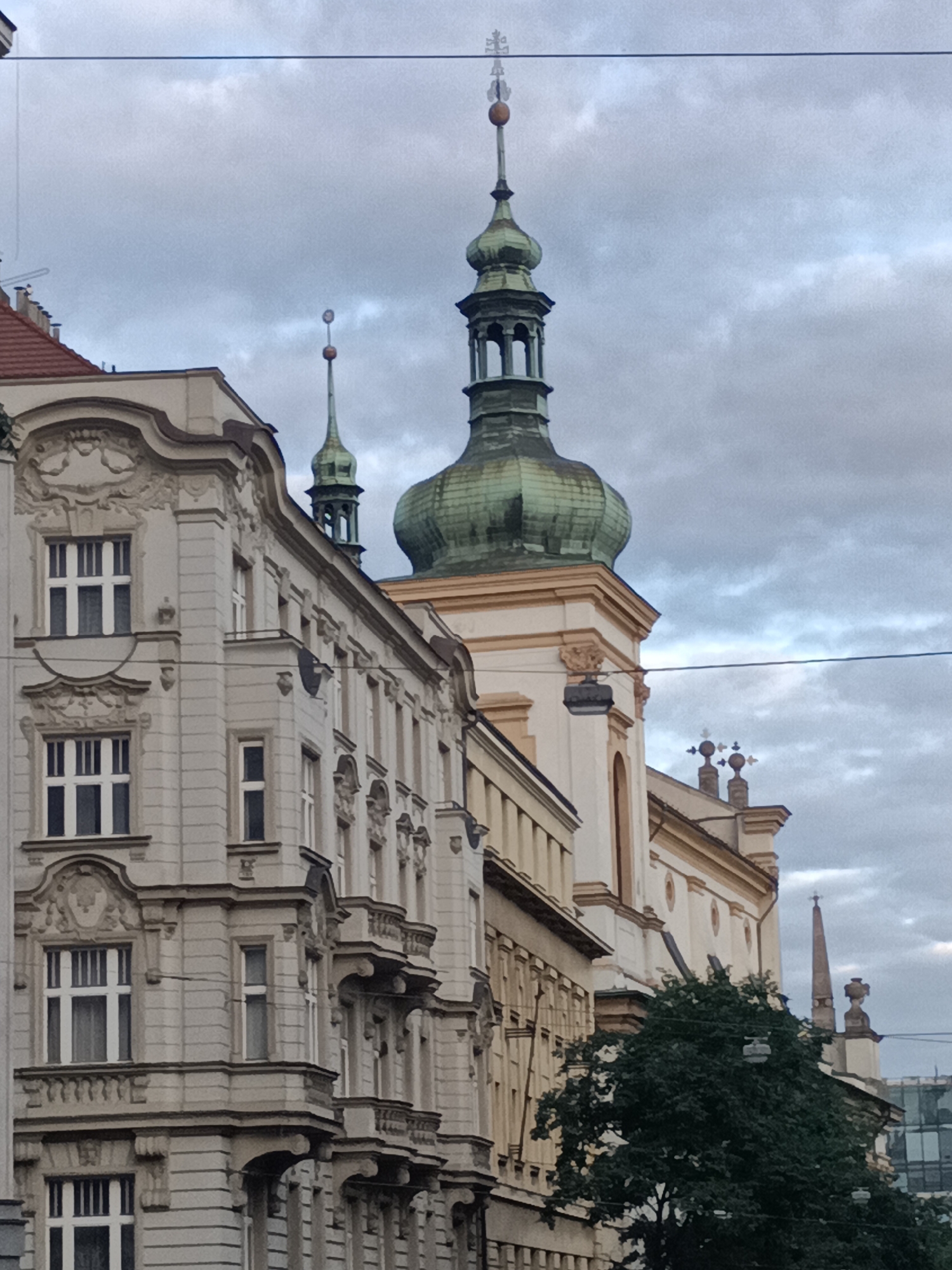 A low-angle, medium shot captures the exterior of ornate European architecture in Prague, Czechia, under a cloudy sky. The foreground features a light-colored, multi-story building with intricate decorative carvings above its windows. The building has a corner facade with a rounded bay on the second story and balconies with decorative railings. Further back, the domed, green-copper roof of a church tower rises above the buildings, topped with a finial. Adjacent to this, another smaller green-copper spire with a finial is visible. In the background, a large dark green tree obscures part of the church structure. Power lines are visible crisscrossing the upper portion of the image. The overall atmosphere is overcast and somewhat subdued.