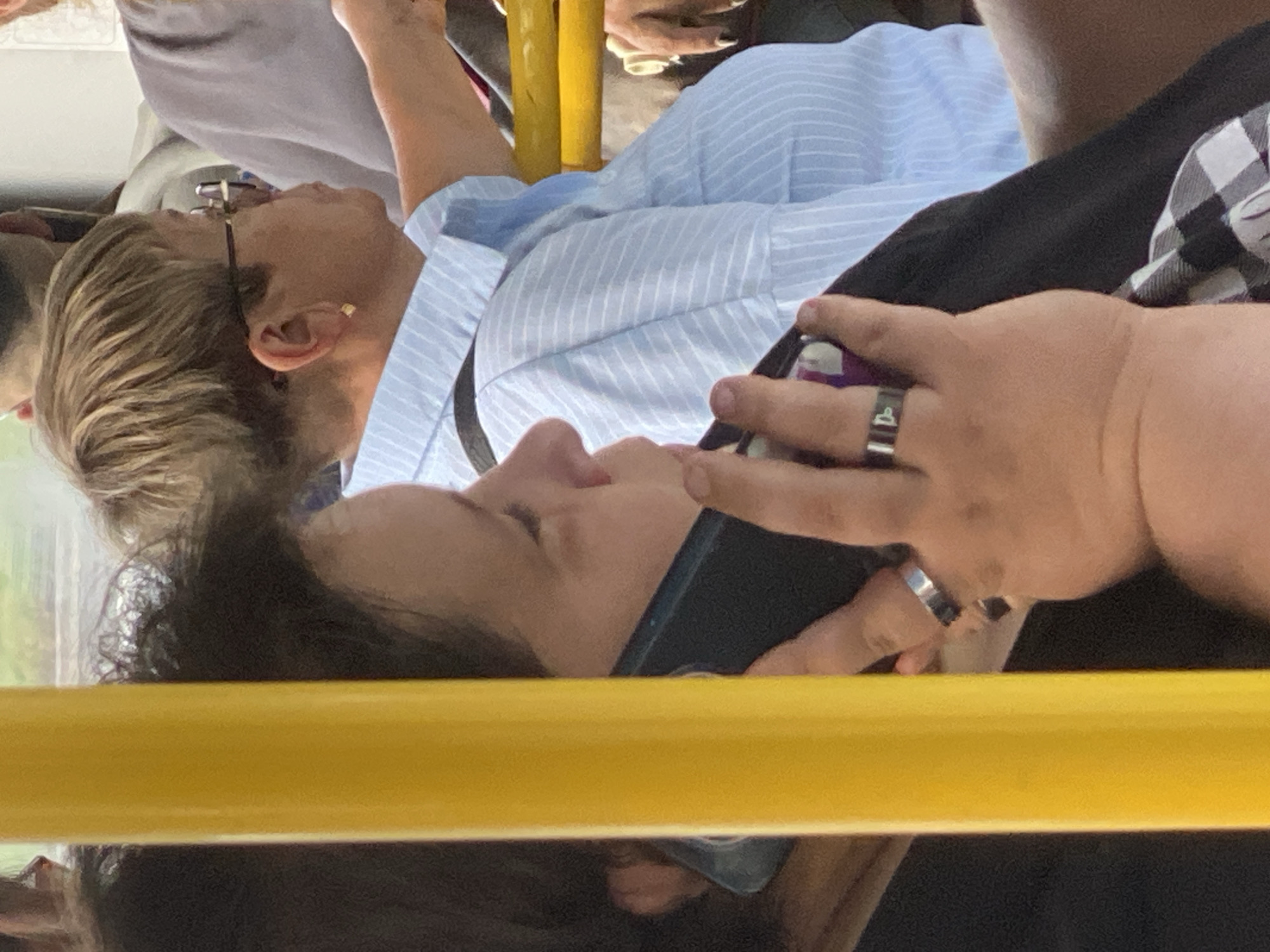 The image captures a close-up, angled view of individuals inside a public transportation vehicle, likely a bus or tram, based on the presence of a yellow pole and yellow grab rails. The focus is on a woman in the foreground, holding a dark-colored smartphone to her ear, suggesting she is engaged in a phone call. She has dark, shoulder-length hair and is wearing a black top. Two rings are visible on her left hand: a silver band on her index finger and another silver ring on her middle finger with an engraved symbol.
Behind her, a woman with short, blonde hair and glasses is visible, wearing a light blue, pinstriped collared shirt. She appears to be looking to her right. Her ear is adorned with a small, square gold earring. Further back, parts of other passengers and the interior of the vehicle can be discerned, including portions of seating and windows. The environment appears to be urban, with daylight visible through the windows, indicating it is daytime. The specific location of Novokuznetsk, Russia cannot be determined from the visual information in the image.