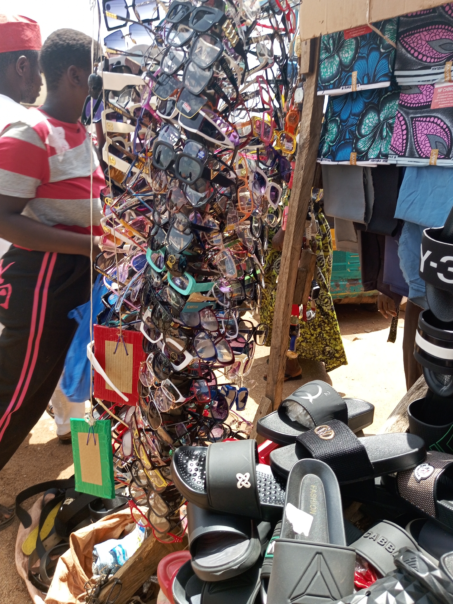 The image depicts an outdoor market scene in Bukuru, Nigeria, likely during the daytime given the bright lighting. The focus is on a stall overflowing with various sunglasses displayed on a rack. The sunglasses are predominantly dark-lensed with a multitude of frame colors and styles, including some with vibrant colored frames like red and teal.
In the foreground, a variety of flip-flops and sandals are piled up, some with distinct brand logos visible, such as "Y-3" and what appears to be "GABBANA." These are primarily black and grey.
To the left of the sunglasses stall, two individuals are partially visible. One person is wearing a red cap and a red and white striped shirt. Another person is wearing black track pants with pink stripes and a striped t-shirt. They appear to be interacting with the stall or browsing.
Behind the sunglasses, a selection of colorful African wax print fabrics are displayed, pinned to a wooden structure. These fabrics feature vibrant patterns in shades of blue, green, pink, and black.
A small, bright green rectangular sign with a brown center is hanging from the sunglasses rack.
The overall impression is a bustling market environment with a focus on retail of fashion accessories. The ground appears to be sandy or dusty, typical of an outdoor market setting. The weather seems to be sunny.
