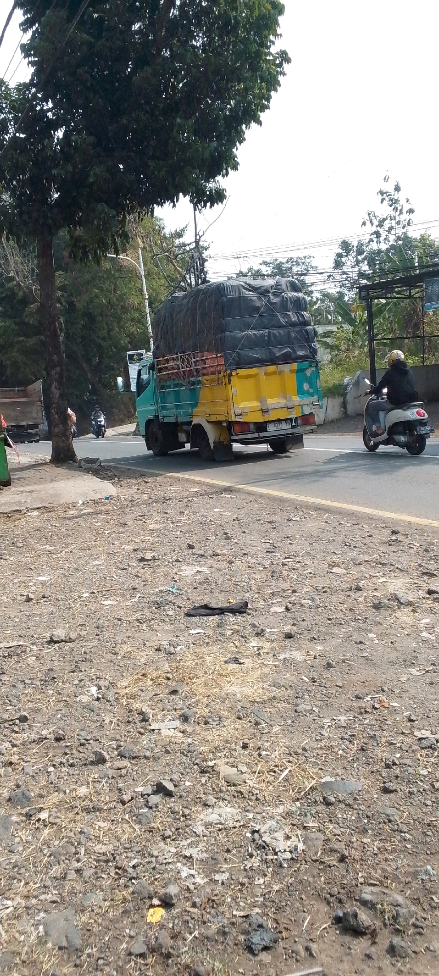 This image depicts a street scene in Cilaku, Indonesia, on a bright, possibly hazy day. A two-toned truck, teal on the front and yellow on the back, is driving on the right side of the paved road. The truck's bed is covered with a large black tarp. To the right of the truck, a person wearing a yellow helmet and a black jacket is riding a white scooter. The road is bordered by a gravel shoulder with some scattered debris. To the left of the road, a large tree with dense green foliage stands next to a narrow sidewalk. Further in the background, there are more trees and some structures, suggesting a semi-urban or roadside environment.