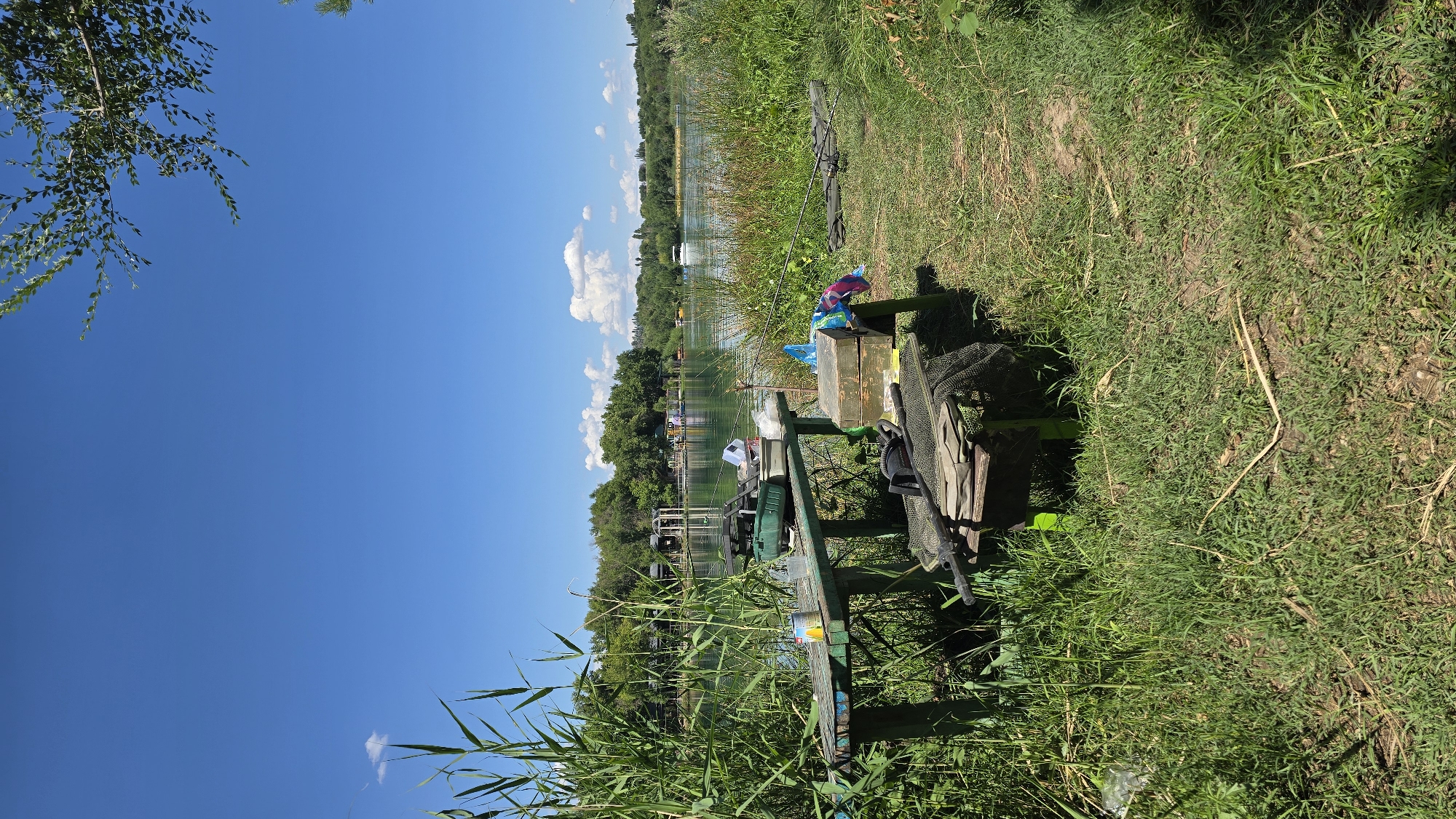 A fishing setup is arranged on a wooden table next to a body of water, likely a lake, on a sunny day in Қарабұлақ, Kazakhstan. Tall reeds and green grass dominate the foreground, with a clear blue sky overhead. The table is laden with fishing gear, including tackle boxes, nets, and what appears to be fishing rods leaning against a nearby stand. A wooden box with colorful cloths draped over it sits on the ground beside the table. In the background, across the water, are trees and structures that suggest a developed recreational area, with a white boat visible on the lake. The scene is peaceful and indicative of outdoor leisure activities.