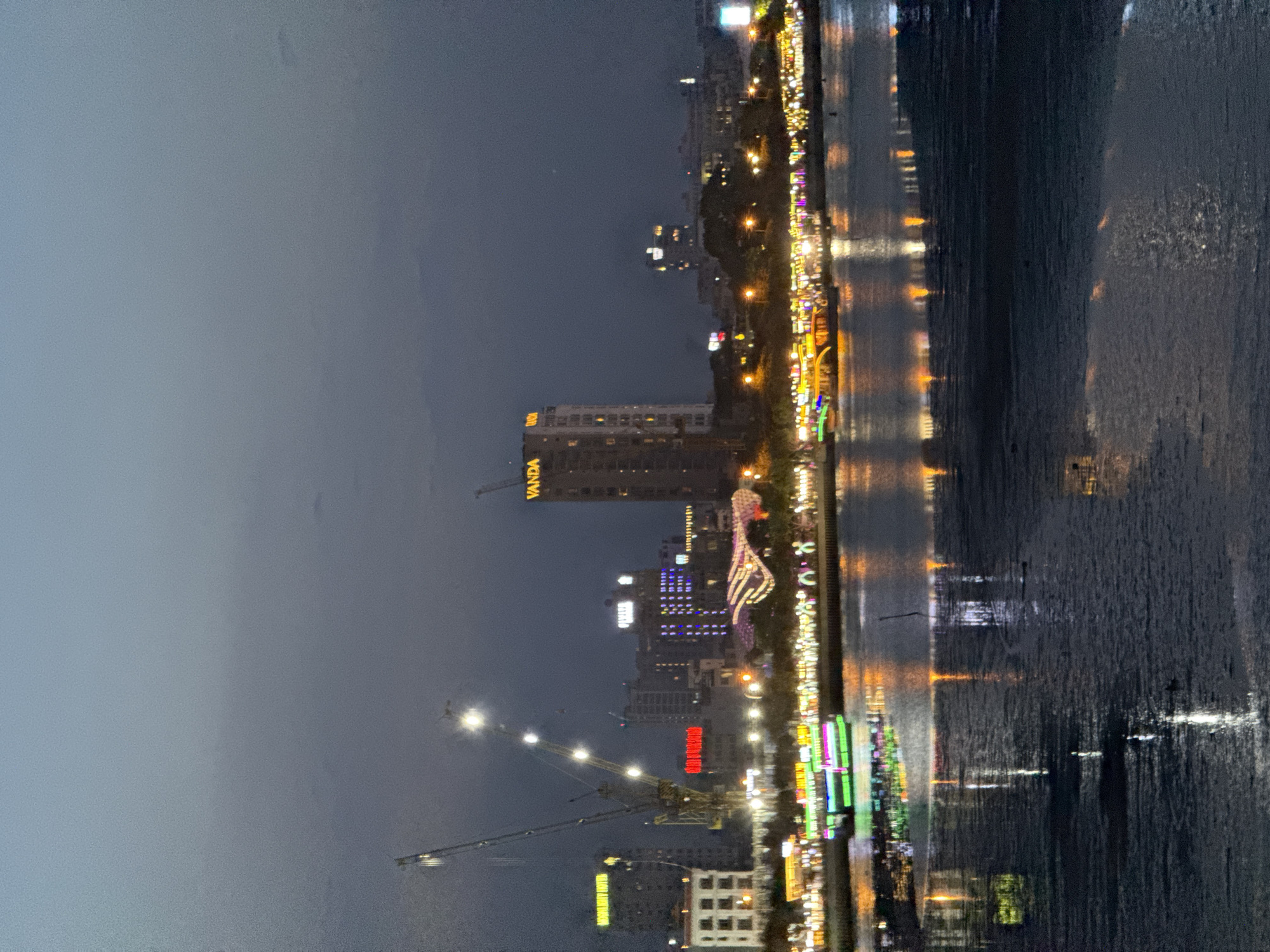 The image shows a waterfront scene in Da Nang, Vietnam, at dusk. The cityscape is illuminated by the lights of buildings and a brightly lit boat on the river. Cranes are visible in the background, suggesting ongoing construction. The water reflects the lights, creating a shimmering effect. The sky is a deep twilight blue, with the faintest hint of clouds. The foreground is dominated by the dark water and its reflections. The scene is calm and serene, with a sense of urban vibrancy and activity.