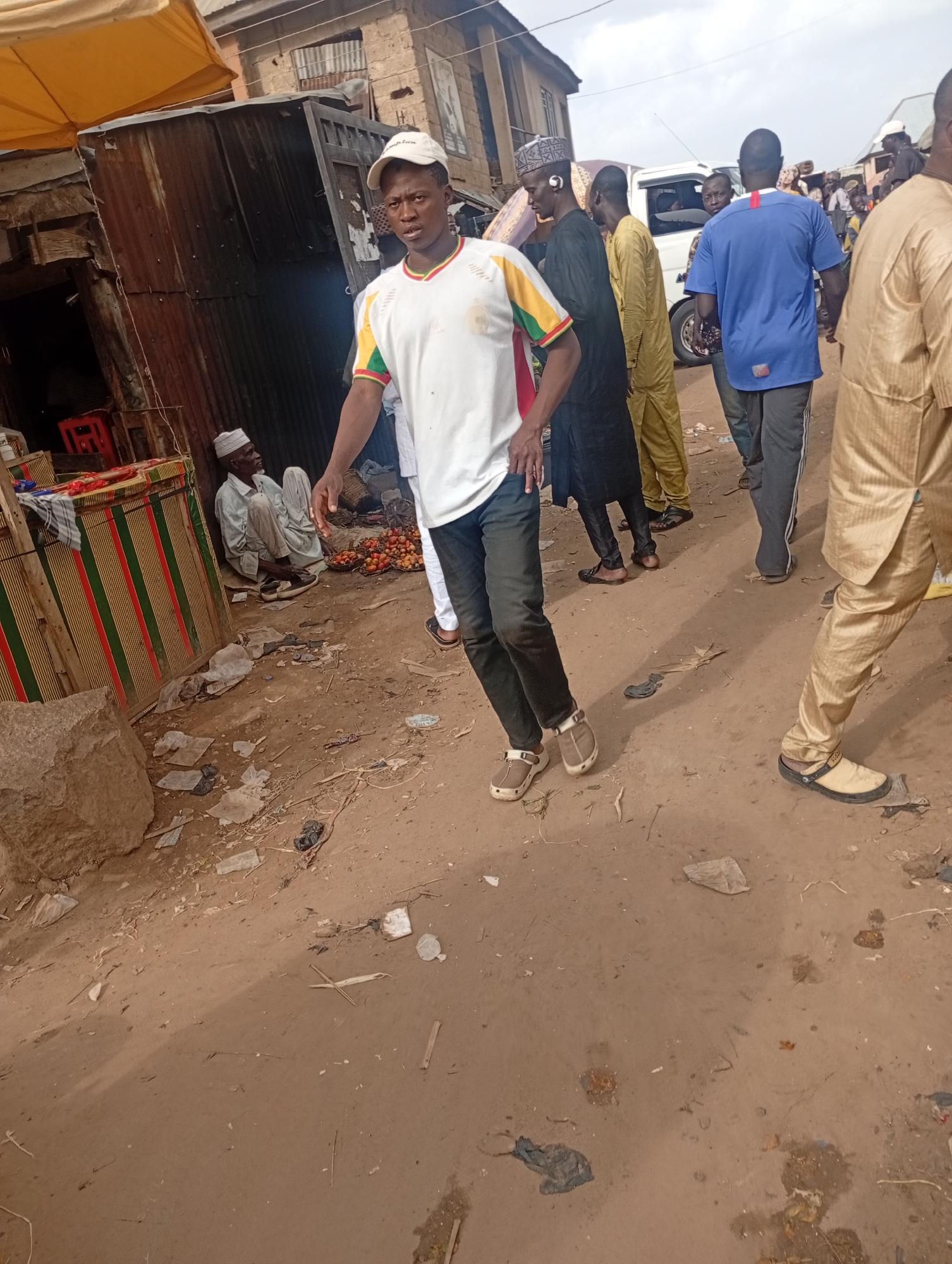 This is a street-level, medium-shot photograph taken in Rafin Par, Nigeria, likely in a market setting. In the foreground, a man wearing a white t-shirt with colored trim and a white baseball cap is walking towards the camera. He is wearing dark pants and light brown slip-on shoes. Behind him, several other people are visible, some walking and some standing.
To the left of the foreground subject, a man in a white cap and traditional light-colored clothing is seated on the ground next to a stall draped with fabric featuring red, green, and white stripes. In front of him, a basket of red fruits, possibly tomatoes, is visible. Further back, a white van is parked. The ground is unpaved and dusty, with scattered debris including plastic bags and other refuse. The background shows buildings with corrugated metal roofs, suggesting a common architectural style in the area. The overall scene depicts a busy, everyday street environment.