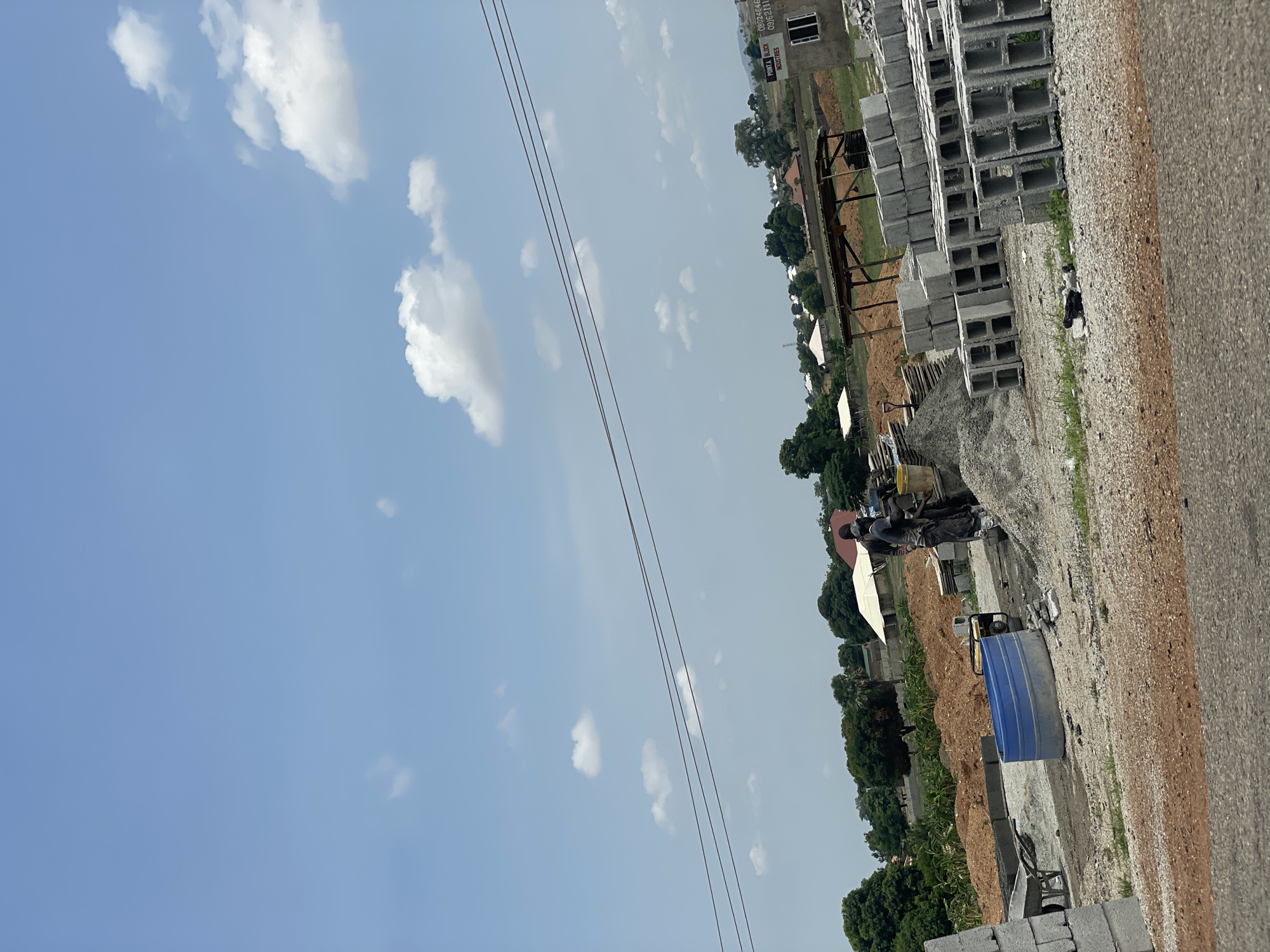 The image depicts an outdoor scene in Kwali, Nigeria. The foreground shows a stack of grey concrete blocks, likely construction materials. A grey asphalt road runs alongside the blocks. Further in the background is construction equipment, piles of gravel, and a blue container. Beyond this, trees and low-lying buildings are visible under a sky with scattered clouds and power lines. There is no indication of any people. The scene implies ongoing construction or industrial activity.