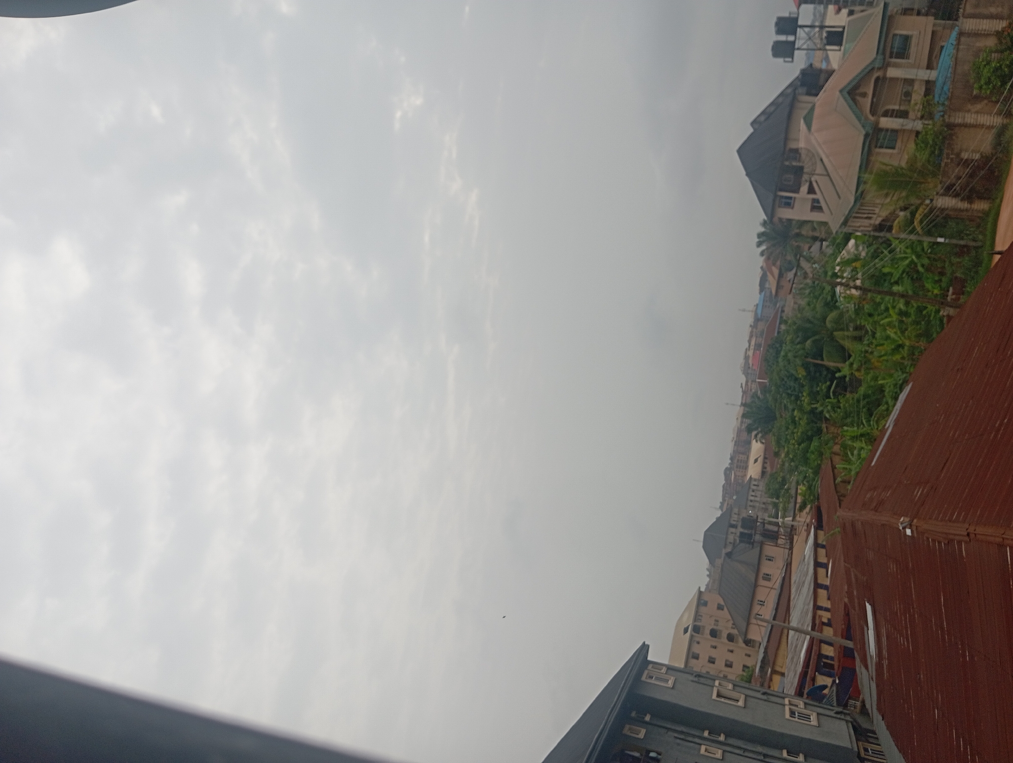 The image shows a cityscape in Nkpor, Nigeria, under an overcast sky. Buildings of varying heights and architectural styles are visible, including residential structures with corrugated metal roofs and taller buildings with multiple stories. Dense green vegetation is interspersed among the buildings. The overall scene suggests a densely populated urban environment with a mix of residential and commercial activity. No people are visible in the image.