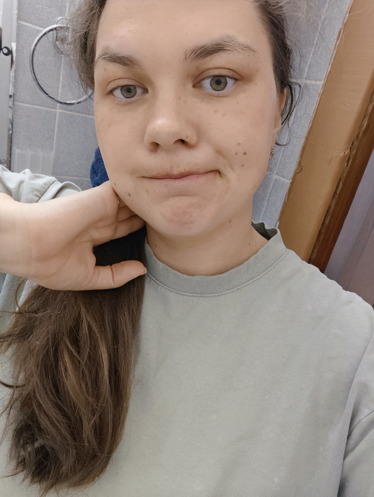 A selfie taken indoors, possibly in a bathroom. A woman with fair skin, light brown hair pulled back, and green eyes is the central subject. She wears a light green t-shirt. Her hand is raised to her neck, holding a dark blue object. The background includes light blue tiled walls, a metal towel rack, and a partially visible wooden door frame. There is no clear indication of the city of Chaikovsky, Russia, in the image.