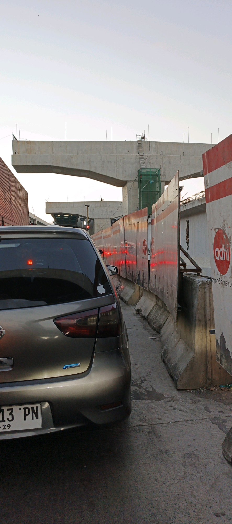 The image shows a street view in Trihanggo, Indonesia, featuring a road under construction. A silver car is parked in the foreground, its rear facing the camera. The car has a license plate reading "13 PN" and a small blue emblem below it. To the right of the car, a construction barrier made of concrete and corrugated metal runs along the road. Red lights are visible on the barrier, likely indicating traffic control or hazard warnings. In the background, a large concrete overpass or bridge structure is under construction, with scaffolding and green netting visible. The sky is overcast with a pale, hazy light. The scene appears to be a traffic jam or a paused moment during construction activities.