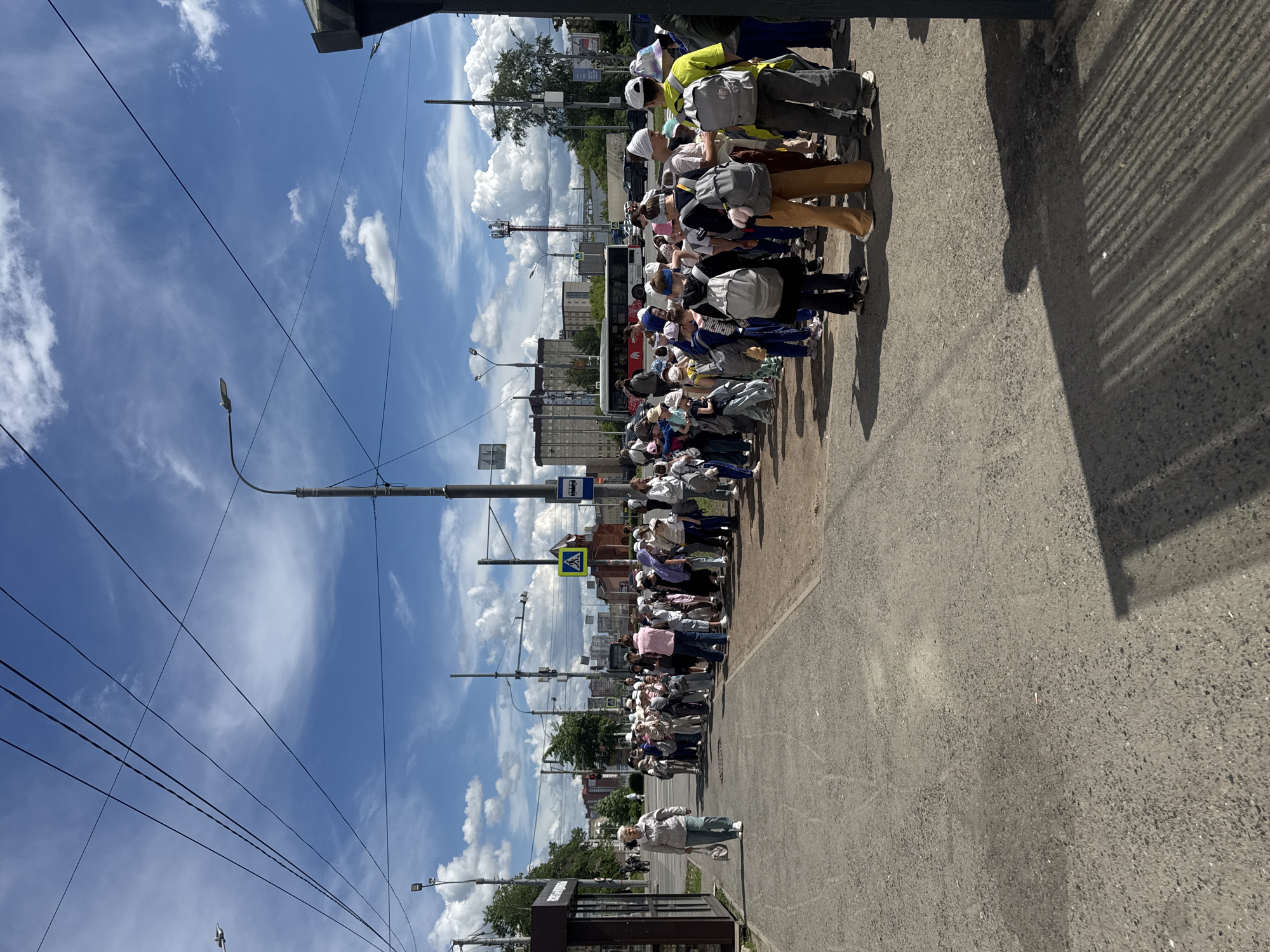 A line of people, the majority of whom are wearing white hats or head coverings, are standing on a street in Perm, Russia. Many are wearing backpacks. The line is on the right side of the road, with a bus visible behind them. A street sign indicating a crosswalk is nearby. A woman is walking near a bus stop, and power lines and street lamps line the opposite side of the street. The sky is blue with scattered clouds. The road is paved and appears to be moderately used.