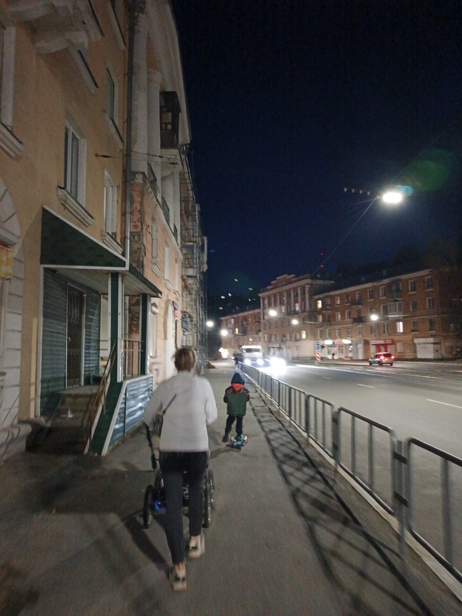 A full-shot, low-angle, nighttime, outdoor image shows a woman walking on a sidewalk in Zlatoust, Russia, pulling a stroller. The woman is wearing a light gray hooded sweatshirt, dark pants, and white sneakers. She is facing away from the camera. A child, wearing a green jacket with orange accents and dark pants, is riding a scooter on the sidewalk ahead of the woman.
The sidewalk is paved and runs alongside a street with traffic. There are streetlights illuminating the area, and several buildings are visible in the background. The buildings appear to be apartment complexes with lit windows. Cars are driving on the street, their headlights creating streaks of light due to the long exposure. A metal railing separates the sidewalk from the street. The overall atmosphere of the image is urban and nocturnal.