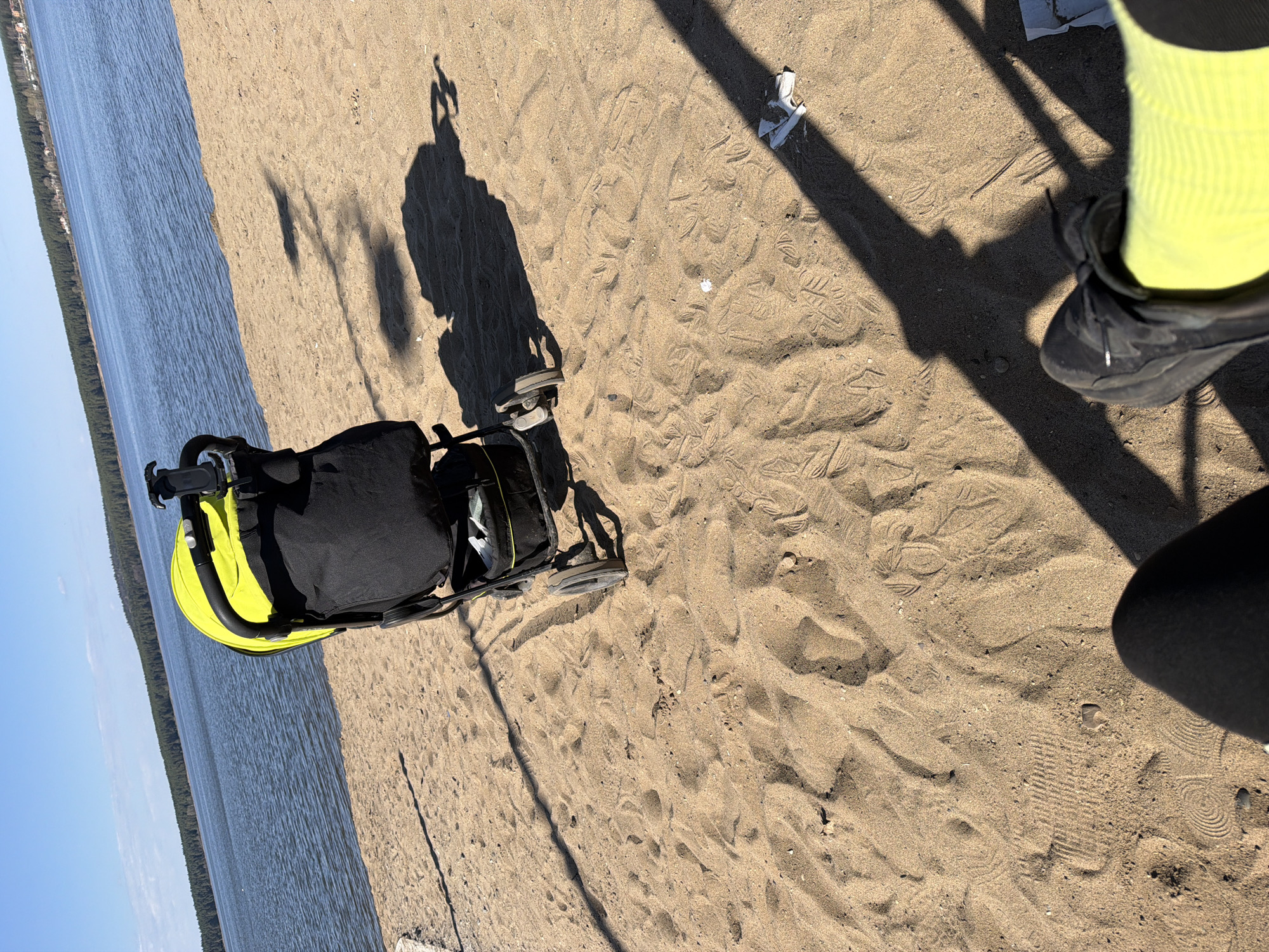 Scene: A sandy area next to a body of water, possibly a beach or shoreline. The location may be in Votkinsk, Russia, based on context.
Subjects/Objects:
1. A black and yellow stroller is positioned on the sand.
2. Footprints are visible in the sand.
3. A pair of feet in dark shoes and light-colored socks are partially visible at the edge of the frame.
4. Shadows of a person and the stroller are cast onto the sand, indicating a light source (likely the sun).
5. Water is visible in the background, with trees lining the far shore.
6. A piece of white material (possibly paper or plastic) lies on the sand.
Actions/Interactions:
-A person is walking or standing near the stroller.
Environment:
-The setting appears to be outdoors on a sunny day.
-The ground is sandy.
-A body of water is present.