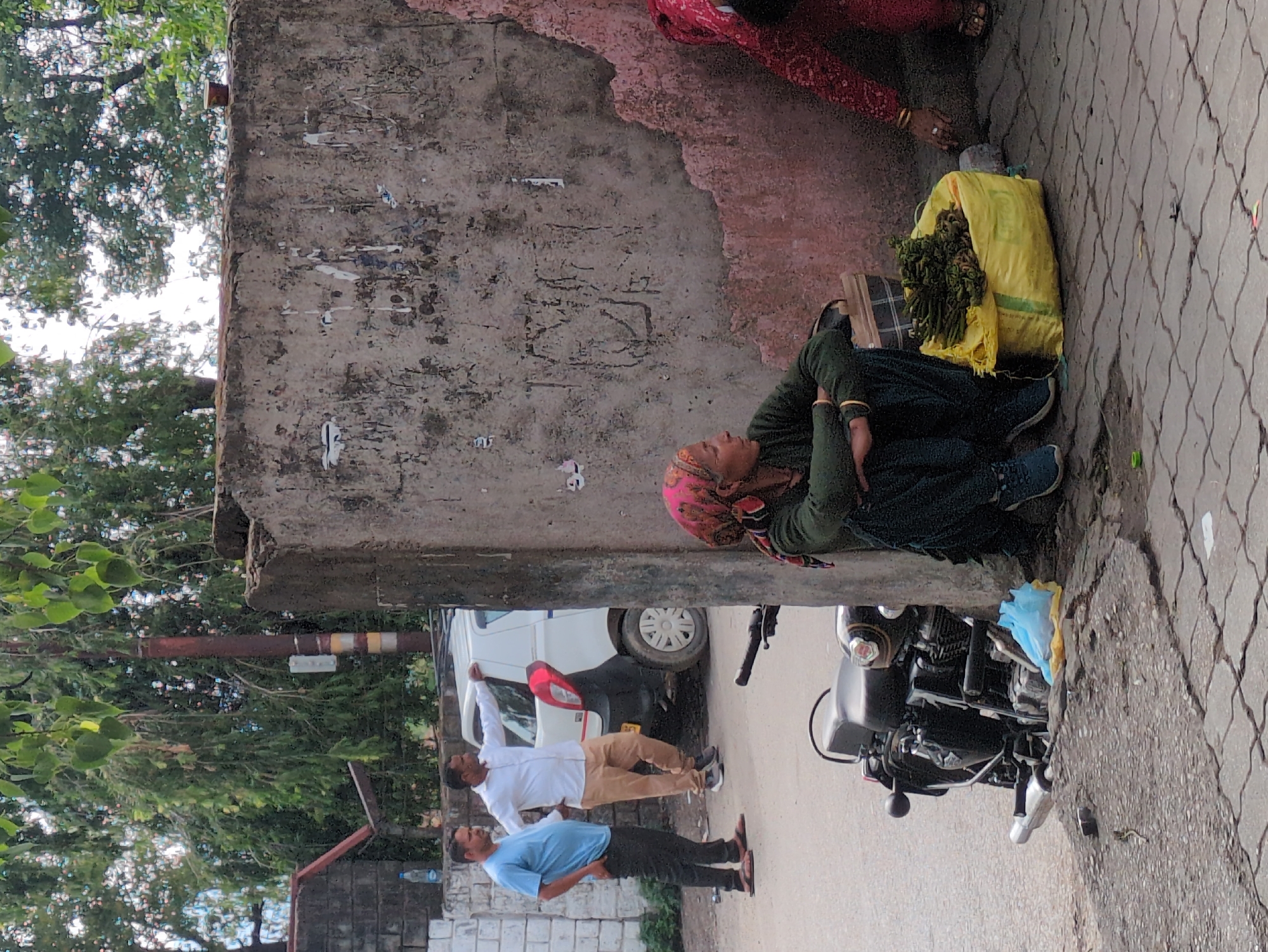 The image depicts an outdoor scene on a paved street, likely in Jhalwan, India, with a textured, weathered concrete wall as a prominent backdrop. Two men, one in a light blue t-shirt and black pants, and the other in a white shirt and tan pants, stand near a white car. The man in the white shirt has his hand resting on the car. In the foreground, a woman wearing a green sweater and dark pants with a pink headscarf is seated on the ground next to a large yellow bag filled with green produce, possibly vegetables. Another person in a red garment is partially visible to the right, reaching towards the yellow bag. A black motorcycle is parked on the left side of the image, near a blue plastic bag. The environment appears to be a street with trees and foliage visible in the background.