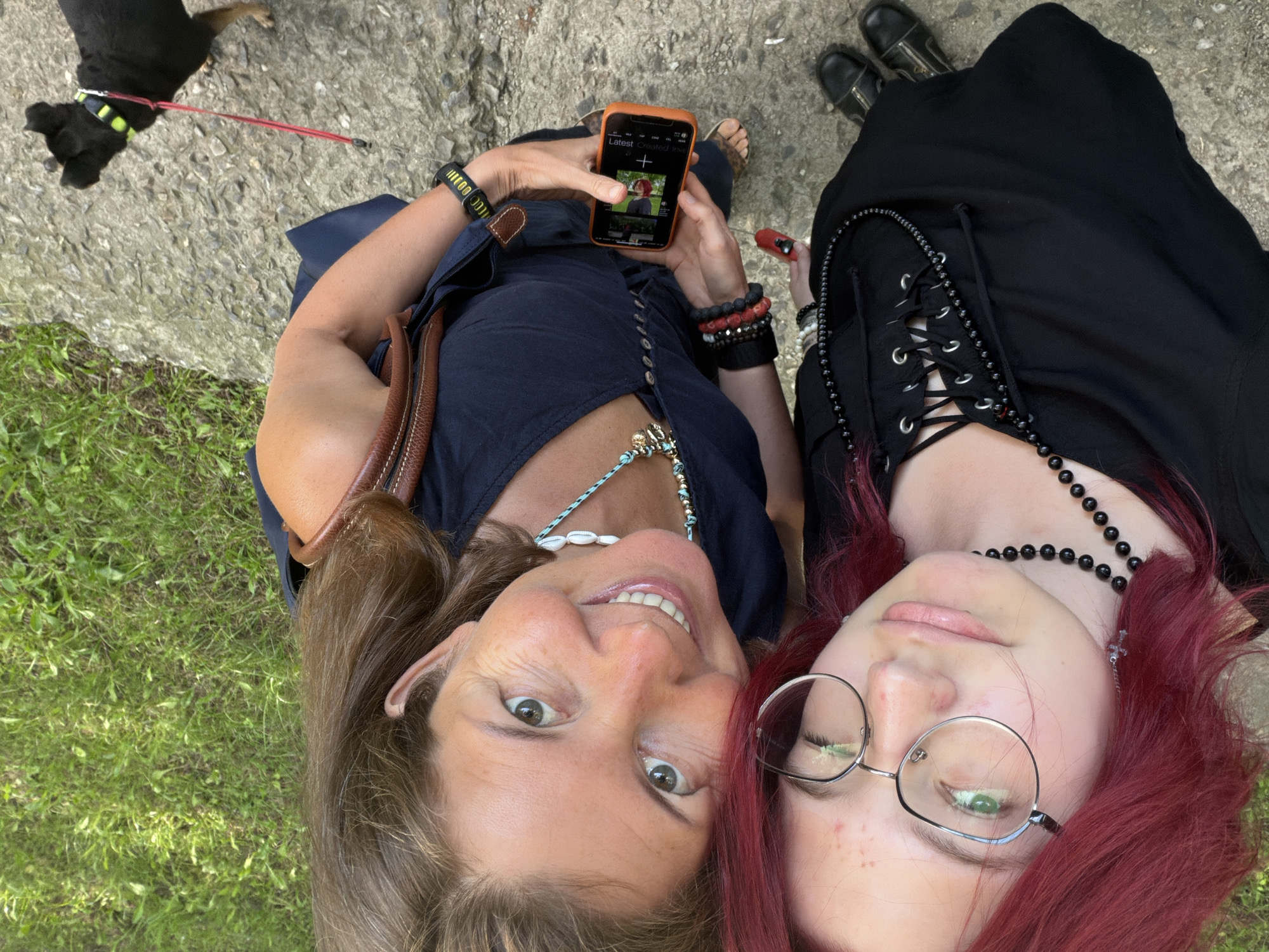 Two individuals, one with vibrant red hair wearing glasses and a black lace-up dress, and another with brown hair wearing a navy blue dress, are posing for a selfie outdoors. The red-haired individual is on the left, and the brown-haired individual is on the right. The latter is holding an orange-cased smartphone, displaying a social media app with a photo of a red apple. Both are wearing necklaces. The red-haired individual has a cross pendant, while the brown-haired individual has a shell necklace. Both are also wearing bracelets. The brown-haired individual is also carrying a brown bag. A small black dog on a red leash is visible in the bottom right corner of the frame, looking to the right. The ground appears to be a mix of concrete and dirt, with a grassy area to the right. The lighting suggests it is daytime. There is no indication of this location being in Chelyabinsk, Russia.