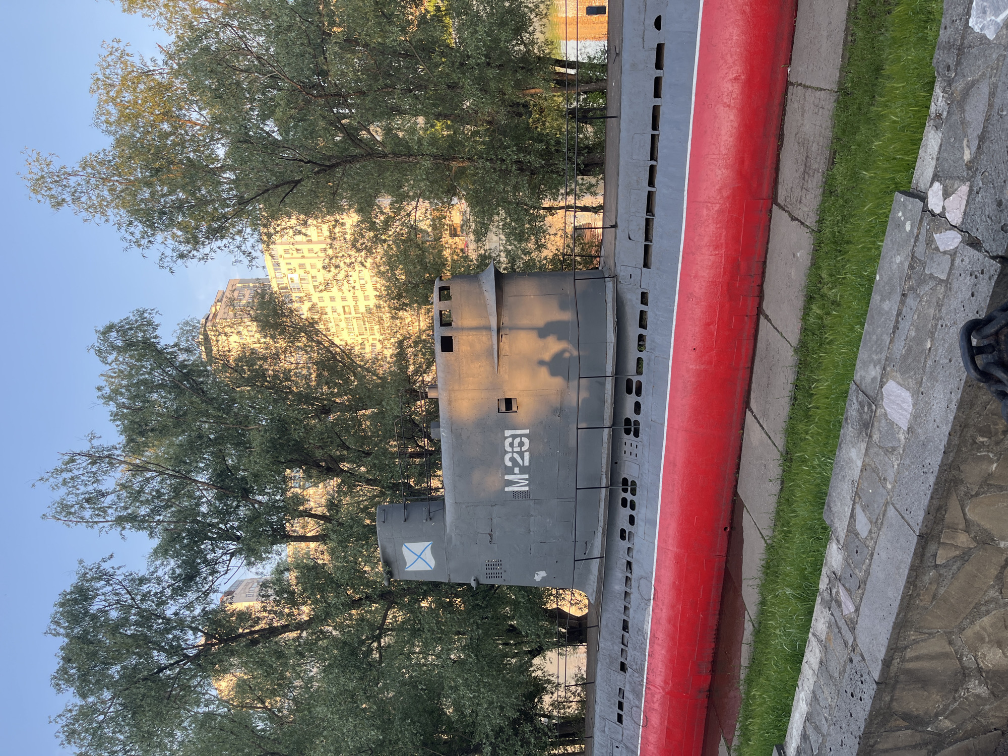 The image shows the Soviet-era M-261 submarine, a museum ship located in Krasnodar, Russia. The submarine is painted grey with a red stripe along its hull. The Cyrillic letters "M-261" are painted on the conning tower. The submarine is positioned on a concrete platform with grass and stone paving around it. Trees are visible in the background, obscuring some of a multi-story building. The sky is light blue.