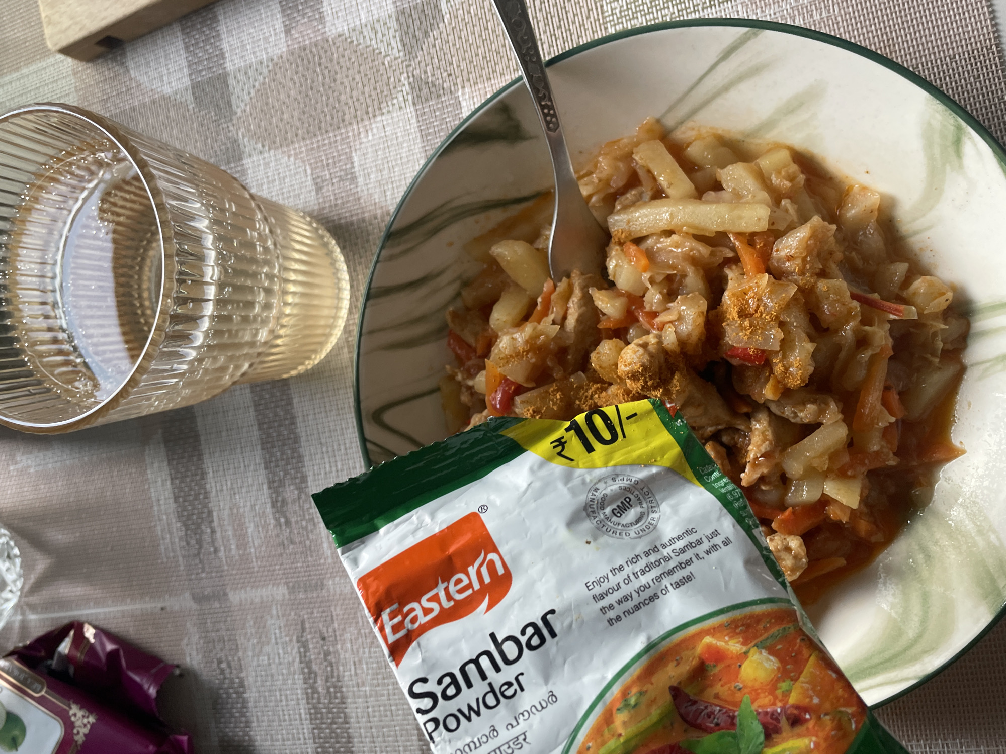 A top-down shot shows a meal of what appears to be stew in a bowl, alongside a glass of water and a packet of Sambar Powder. The meal is served in a light-colored bowl with a green rim, and a fork is resting in the food. The Sambar Powder packet is colorful, with a picture of the dish on it, and the words "Eastern Sambar Powder" are clearly visible. The packet also displays a price of "₹10/-" and a GMP certification. The setting appears to be a table covered with a checkered tablecloth, and a wooden object is visible in the top left corner, possibly a cutting board. The lighting suggests it is daytime. The overall impression is of a home-cooked meal being prepared or enjoyed.