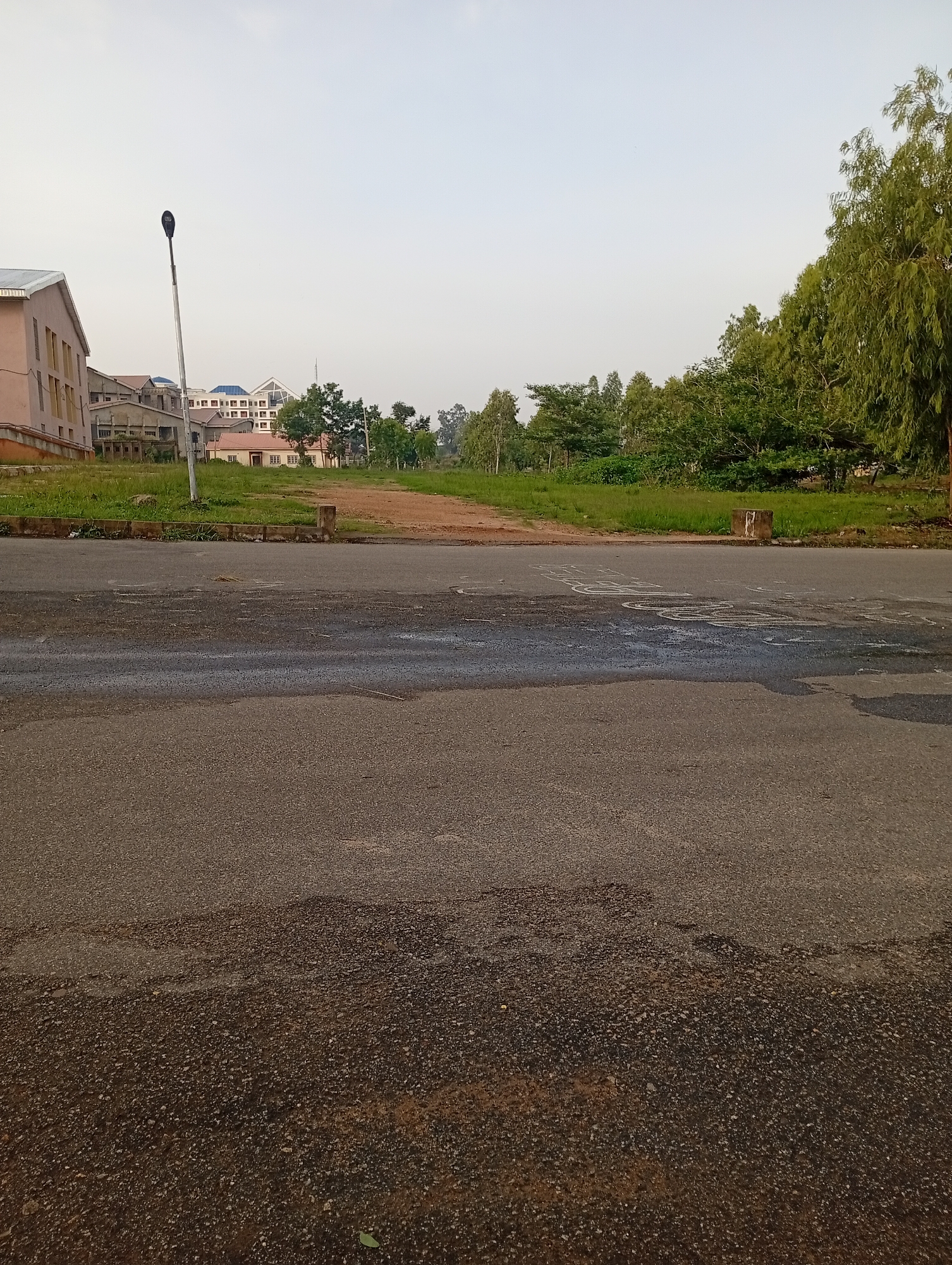 The image depicts an outdoor scene with a focus on a paved road in the foreground, leading towards a grassy area with buildings and trees in the background. The road surface appears to be asphalt, with some patches of wetness and evidence of traffic markings, possibly chalk drawings or faded paint.
In the middle ground, a dirt path or unpaved road ascends gently into the distance, flanked by green grass and vegetation. A tall, modern streetlight stands on the left side of the paved road, near the edge of the grassy area.
Beyond the grassy area, several buildings are visible. On the left, a multi-story building with a light pink facade and numerous windows is prominent. Further back, other structures with lighter-colored walls and tiled roofs can be seen, suggesting a campus or residential area. The architecture appears contemporary.
A line of trees, varying in size and density, forms a backdrop to the buildings, extending across the mid-ground and into the right side of the frame. The foliage is lush and green, indicating a temperate climate. The sky above is a pale, hazy blue, with no distinct clouds.
The overall impression is one of an urban or semi-urban environment, possibly on the outskirts of a city or within a developed area characterized by both modern infrastructure and natural elements. The lighting suggests it is daytime, likely morning or late afternoon, given the soft light and long shadows are not prominent. The scene is devoid of people or any observable activity, presenting a tranquil yet static view of the location. The presence of the streetlight, paved road, and multi-story buildings suggests an established infrastructure.
Considering the visual cues such as the architectural style of the buildings, the type of vegetation, and the general landscape, this scene could plausibly be located in a developed area within Jos, Nigeria, known for its cooler climate and greenery.