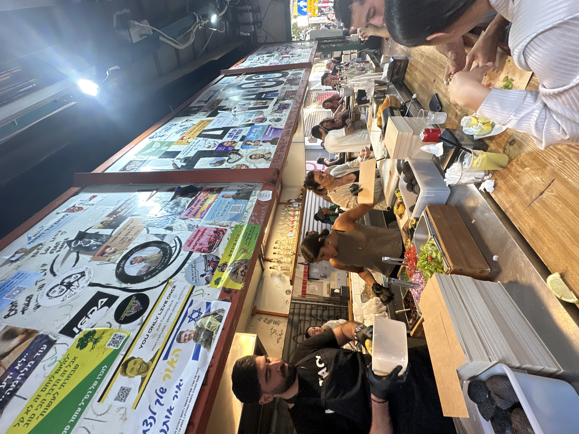 The scene is a bustling outdoor eatery in Tel-Aviv, Israel. A long counter is lined with customers eating and drinking, while behind it, food vendors are preparing meals. The counter is wooden, with condiments, drinks, and prepared dishes. The vendors, wearing aprons and gloves, work with fresh vegetables, avocados, and prepared ingredients. Behind them is a collage of stickers, posters, and memorabilia covering the wall, including images of soldiers, phrases in Hebrew, and English slogans. The atmosphere suggests a lively and casual dining experience.