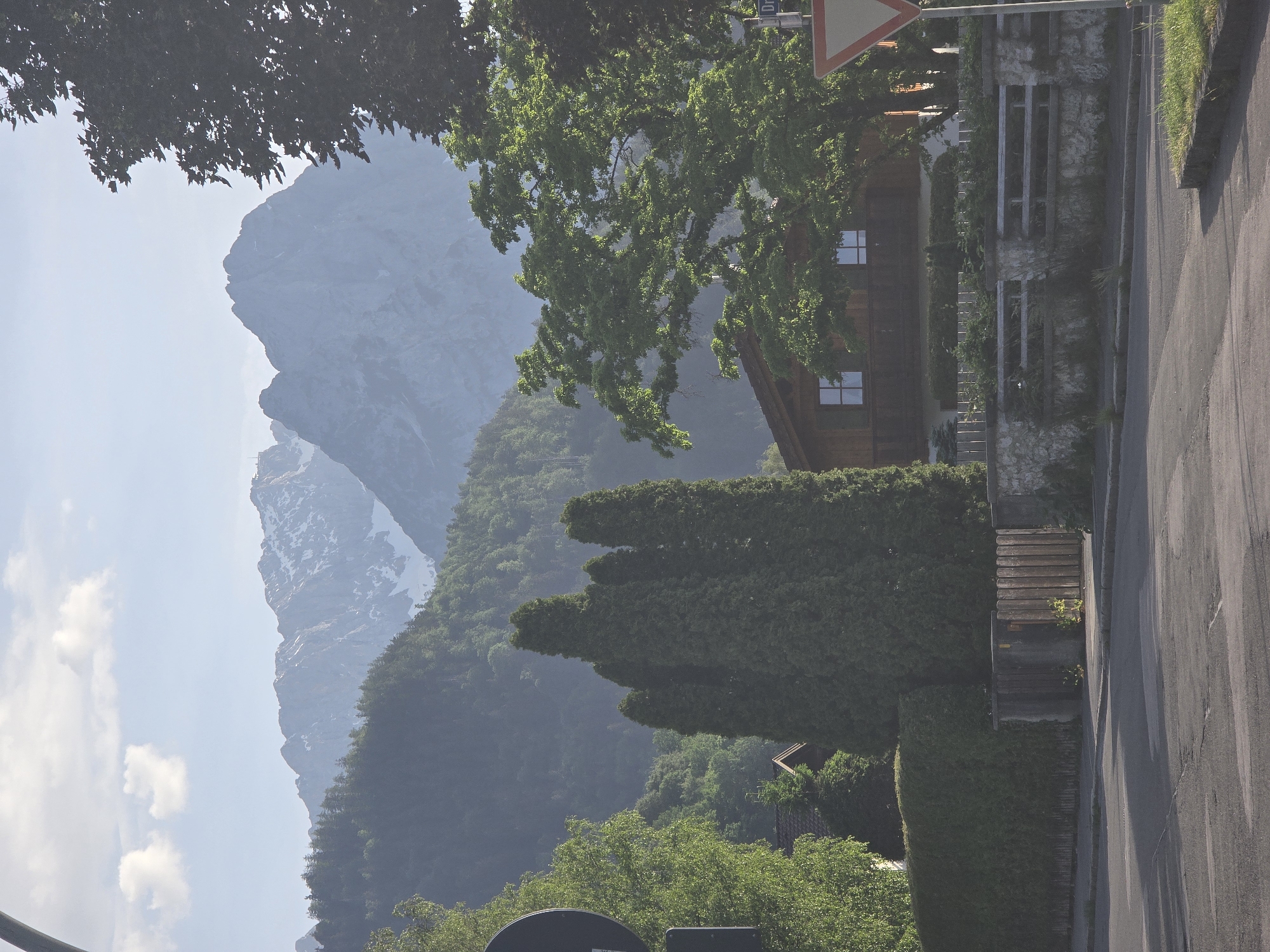 The image depicts a mountain view in Garmisch-Partenkirchen, Germany, framed by trees and vegetation along a street. The road surface is asphalt. A multi-story wooden house stands on the right, partly obscured by tall, shaped hedges. A stone wall is visible beside the house. A mountain range looms in the background, its peaks partially covered with snow. The sky is light blue with some clouds.
