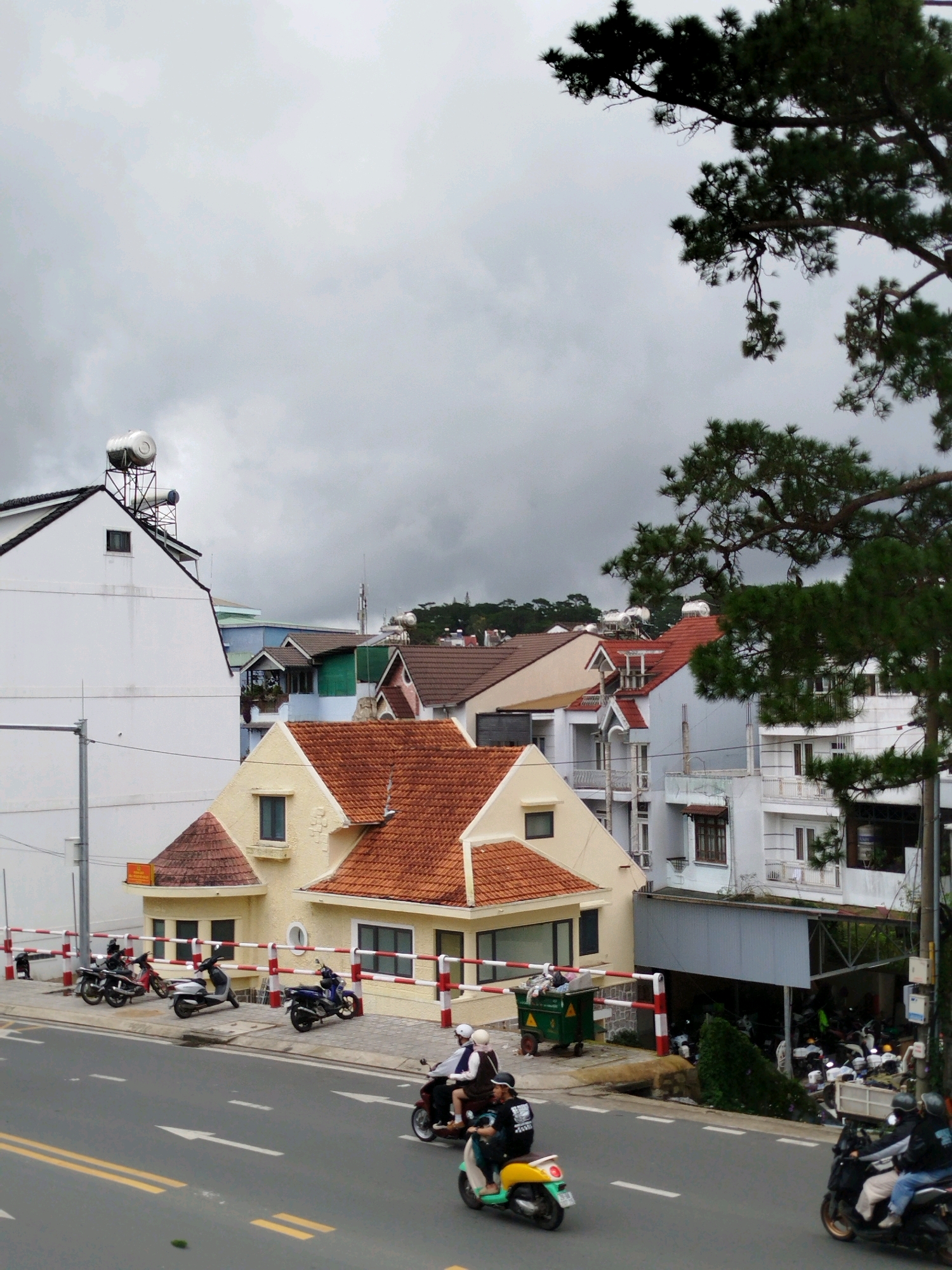 The image depicts a street scene in Da Lat, Vietnam, under a cloudy sky. The foreground shows a road with white and yellow lane markings. Several motorbikes are visible on the road and parked along the sidewalk. Two people are riding a motorbike in the foreground, one wearing a helmet and a dark jacket, and the other a helmet and a light-colored jacket. A second motorbike with two riders, both wearing helmets, is also in the foreground.
Behind the road, a sidewalk area is lined with a red and white striped barrier. Parked motorbikes are visible on this sidewalk. Beyond the sidewalk, there are several buildings with terracotta tiled roofs. A distinctive yellow building with a red-tiled roof and small windows is prominent. To the left of this yellow building is a larger white building with a dark roof. Further in the background, more buildings with various colored roofs are visible, along with trees and hills in the distance. A large green dumpster is positioned on the sidewalk near the yellow building, with a person sitting on it.
The overall environment suggests an urban setting with a mix of residential and commercial architecture. The presence of numerous motorbikes and the style of the buildings are characteristic of Vietnamese cities, and specifically Da Lat, known for its French colonial architecture and cooler climate. The cloudy sky indicates potentially overcast or rainy weather.