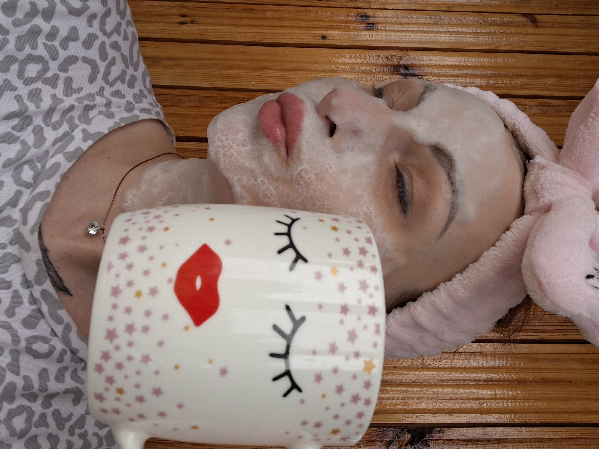 A woman with a pink bunny-ear headband and a white facial mask applied is holding a mug with a stylized face design in front of her face. Her eyes are closed, and her lips are pursed. She is wearing a grey and white leopard-print t-shirt. The background consists of vertical wooden planks. The image appears to have been taken outdoors, possibly on a porch or deck, as suggested by the wooden background and natural lighting.