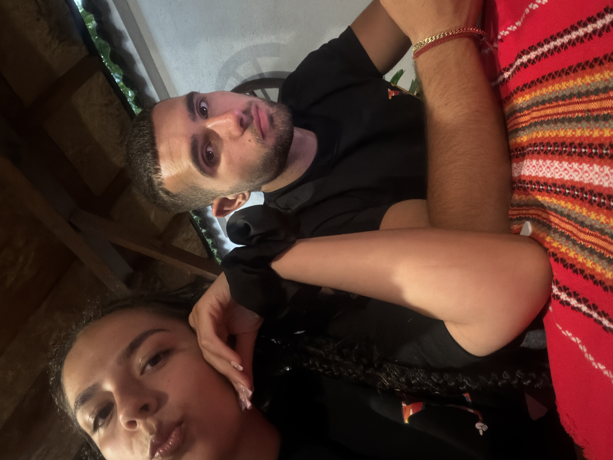 The image shows a close-up, head-and-shoulders portrait of a young man and a young woman sitting at a table. The woman is on the left, with her head resting on her hand and her face turned towards the camera. She has dark curly hair, prominent eyebrows, and appears to be pouting her lips. She is wearing a black top. The man is on the right, looking directly at the camera with a neutral expression. He has short, dark hair, a beard, and is wearing a black t-shirt. He is wearing a gold chain bracelet on his left wrist. They are seated at a table covered with a red tablecloth that has a colorful, striped pattern. The background appears to be an indoor or covered outdoor setting, with wooden beams visible overhead and a white wall behind the man. There is a hint of greenery and what looks like a rustic wooden wheel decoration on the wall. The image seems to have been taken in a casual setting, possibly a restaurant or a home. The location is identified as being in the city of Gabrovo, Bulgaria.