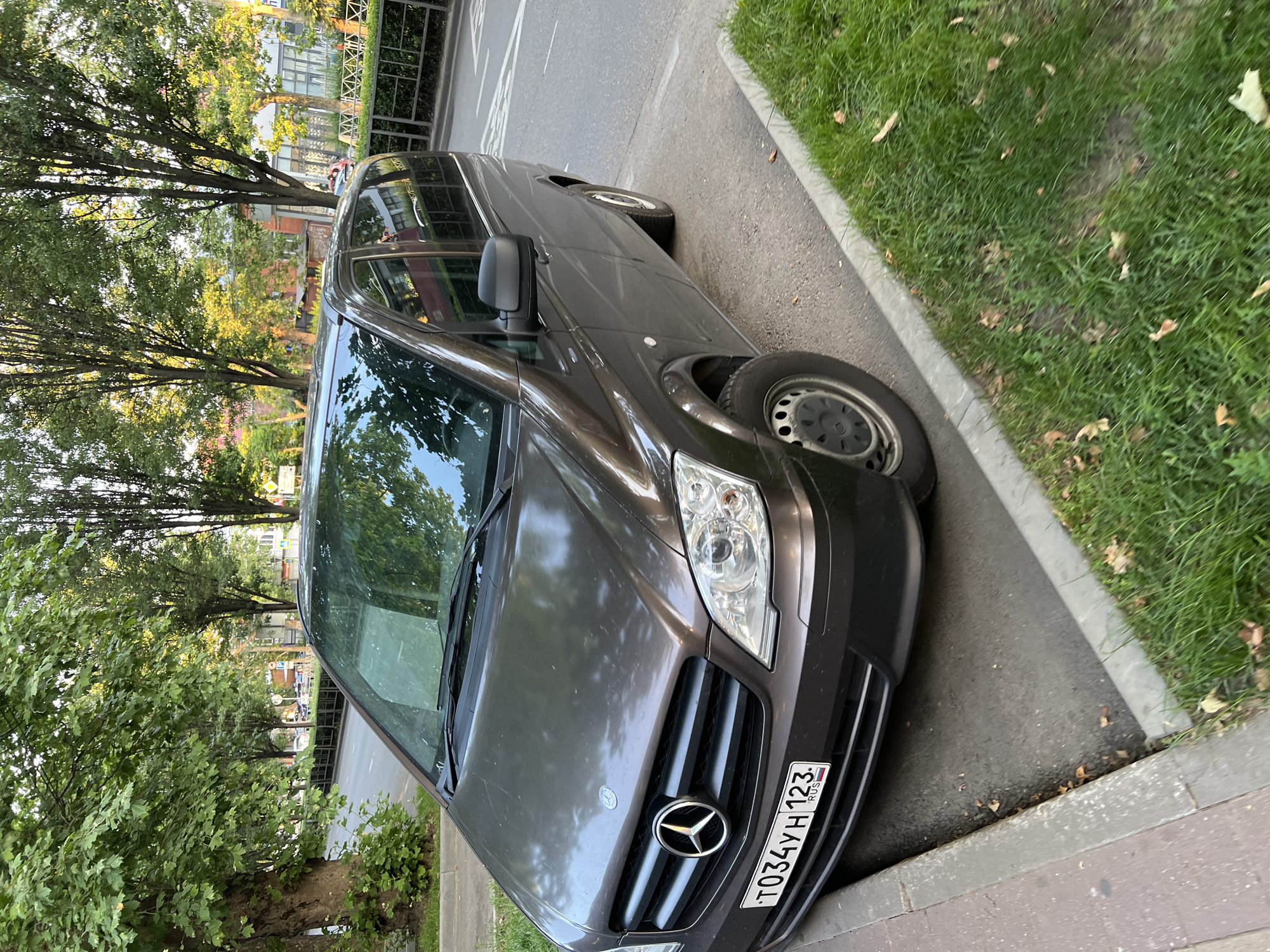 This media file shows a dark brown Mercedes-Benz Vito van parked on the side of a street in Krasnodar, Russia. The van is positioned at an angle, with its front slightly turned towards the camera. The license plate is visible and reads "T034YH 123".
The scene is outdoors, with a paved road on one side of the van and a grassy area with a concrete curb on the other. Mature trees with green foliage line the street, casting shadows on the van and the road. In the background, buildings and more trees are visible, suggesting an urban or suburban environment. The lighting indicates it is daytime, likely late afternoon or early evening, as there is a warm glow on the leaves of the trees. The weather appears to be clear and pleasant.
There are no people prominently visible in the frame. The focus is on the parked van and its surroundings. The van itself is clean and appears to be in good condition. The overall impression is a calm, everyday scene of a vehicle parked on a tree-lined street.