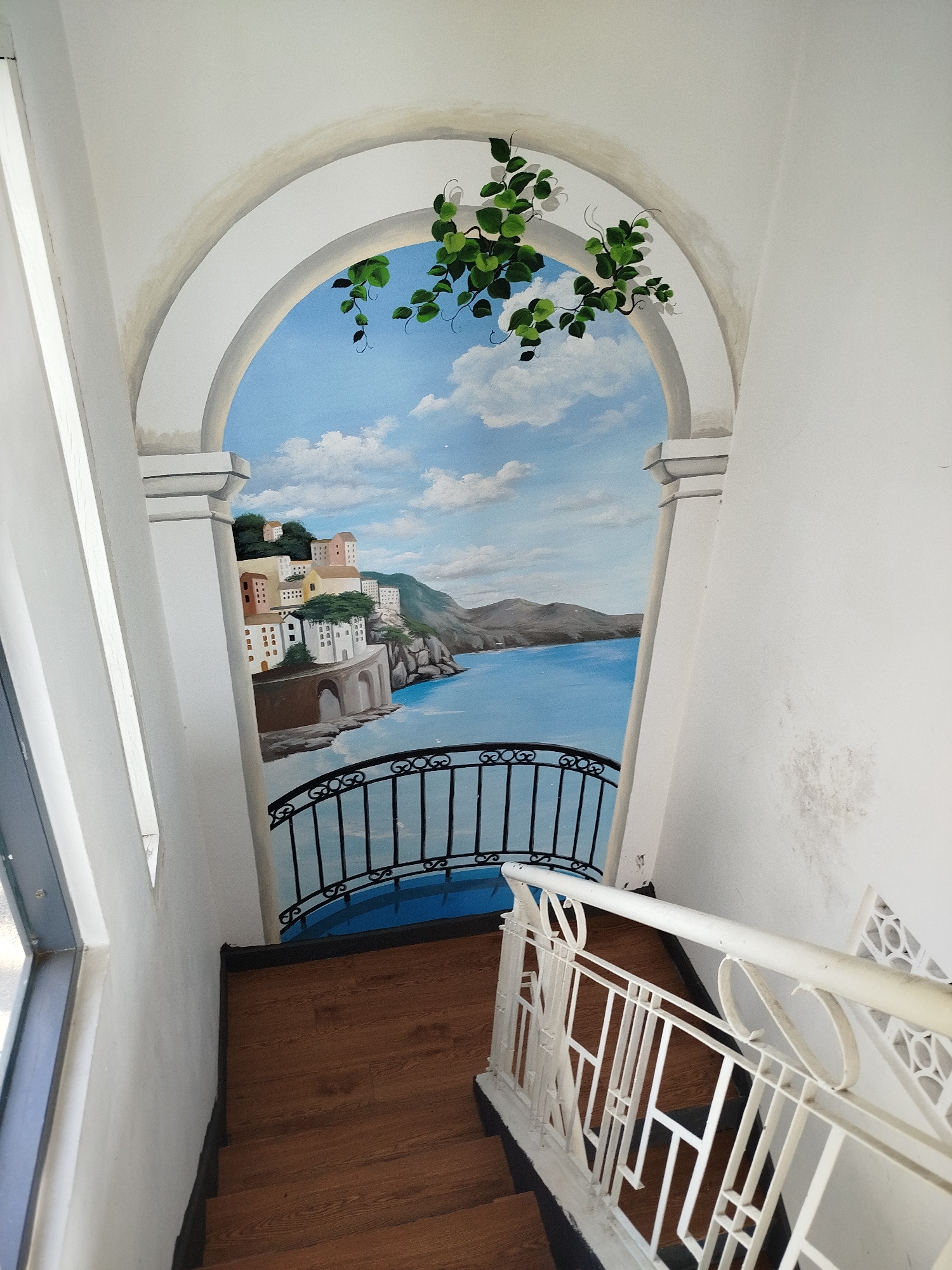 A stairwell features a trompe l'oeil painting of a Mediterranean coastal scene. The painting depicts a view through an archway, framed by architectural elements and adorned with painted foliage. The scene includes buildings on a hillside overlooking a blue sea, a black wrought-iron balcony railing, and a blue platform in the foreground. The stairwell itself has wooden steps and white railings with geometric patterns. The walls are white, with a window on the left and some staining on the right. The scene suggests an attempt to create a scenic illusion within the stairwell. There is no confirmed connection to Da Lat, Vietnam.