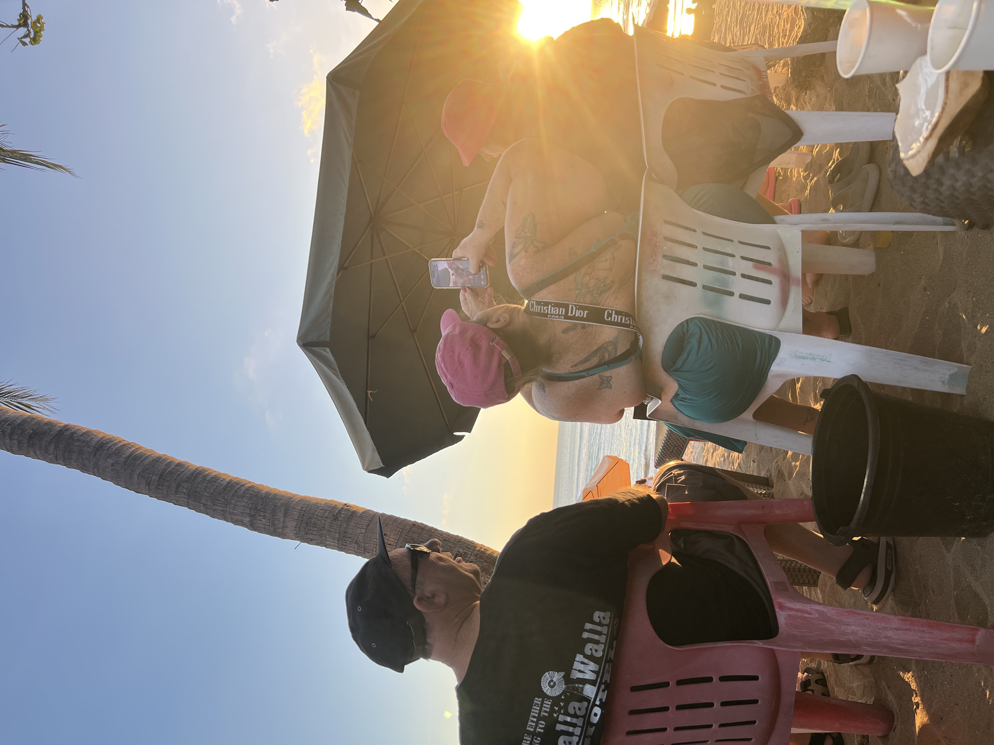 Three individuals are seated on chairs on a sandy beach in Kuta, Indonesia, with the ocean and a sunset visible in the background. A palm tree trunk is on the left side of the frame, and an umbrella is positioned above and behind the seated individuals. The person on the left, wearing a black t-shirt and a cap, is looking to the right. The person in the middle, wearing a pink cap and a green top, is holding a smartphone and taking a picture. The person on the right, wearing a red cap, is also looking towards the sunset. Several cups are on a table or the ground in the foreground. The sun is low in the sky, casting a warm glow and lens flare across the scene.