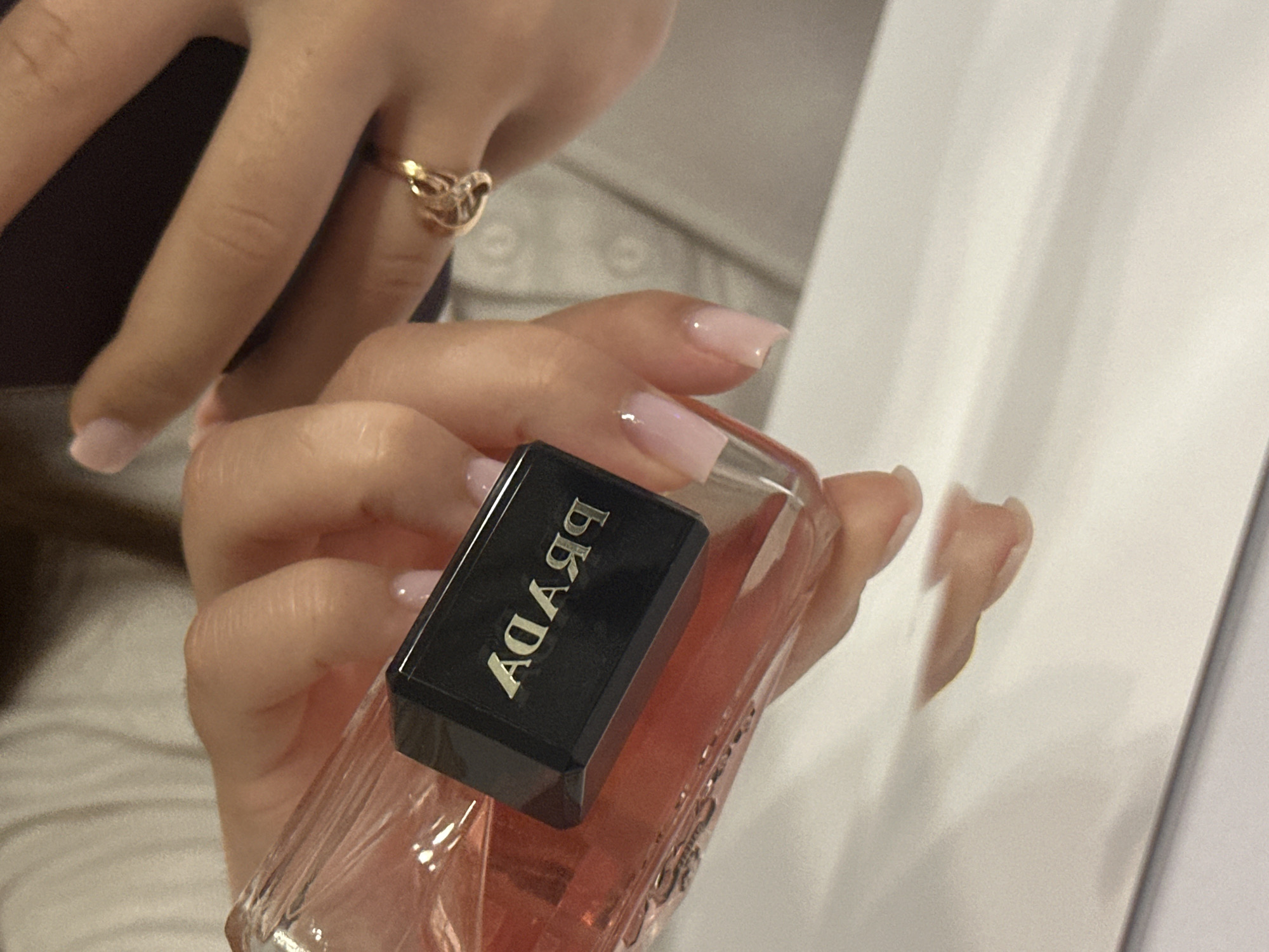 A close-up, downward-angled shot features two hands interacting with objects over a highly reflective, light-colored surface. The left hand, with light pink polished nails, securely grips a clear glass perfume bottle containing a pink liquid. The bottle's black, rectangular cap displays the brand name 'PRADA' embossed in silver, which appears reversed ("ADAR9") to the viewer due to the object's orientation. Beneath the bottle, a distinct reflection of the bottle and the gripping fingers is visible on the surface. To the right, a second hand, identified as the right hand, partially holds a dark-colored smartphone. This hand also exhibits light pink polished nails, with the ring finger adorned by a gold-toned ring featuring an intricate, interwoven design. The background is softly blurred, showing folds of light-colored fabric, suggesting clothing or upholstery. The scene is illuminated by soft, even lighting.