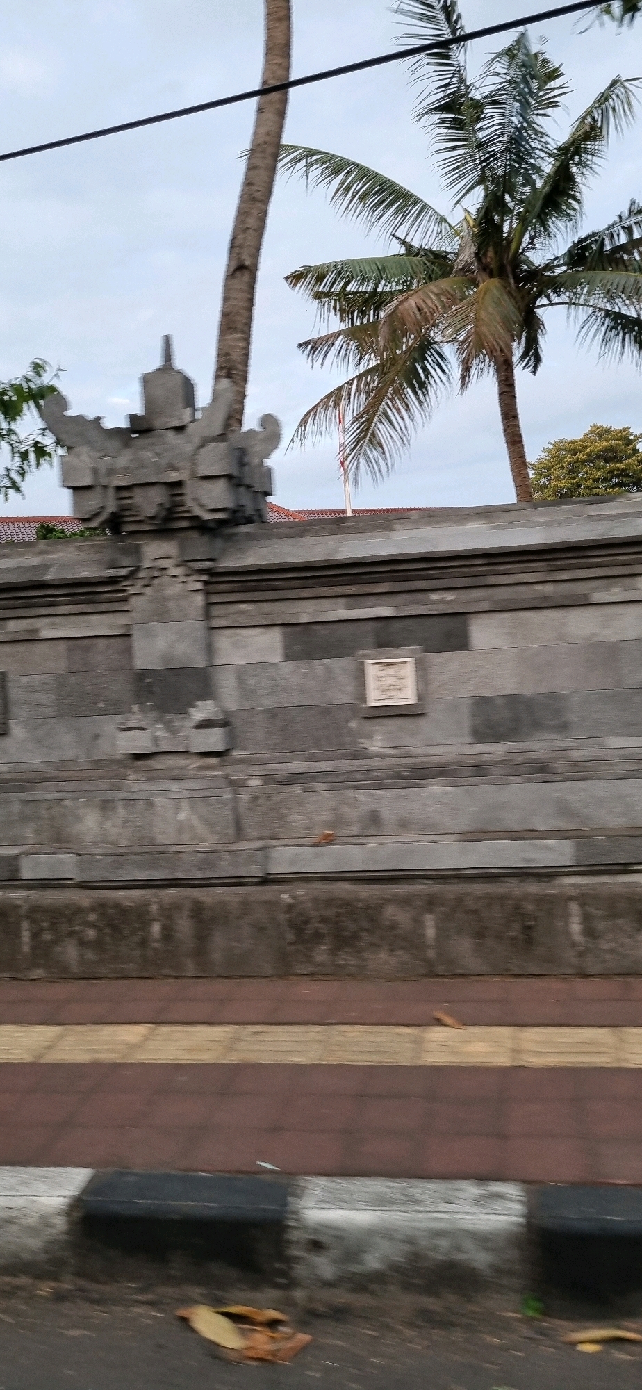 The image shows a stone wall with decorative elements, likely part of a temple or traditional building, in Denpasar, Indonesia. Two palm trees are visible behind the wall, with a cloudy sky above. The wall is constructed from large, grey stone blocks and features intricate carvings at the top. A small, light-colored plaque is mounted on the wall. In the foreground, there is a sidewalk with patterned paving stones in red and yellow, and a curb with a black and white stripe. The ground is paved with asphalt, and a few fallen leaves are scattered on it. No people are visible in the scene. The overall impression is of a roadside view of a culturally significant structure.