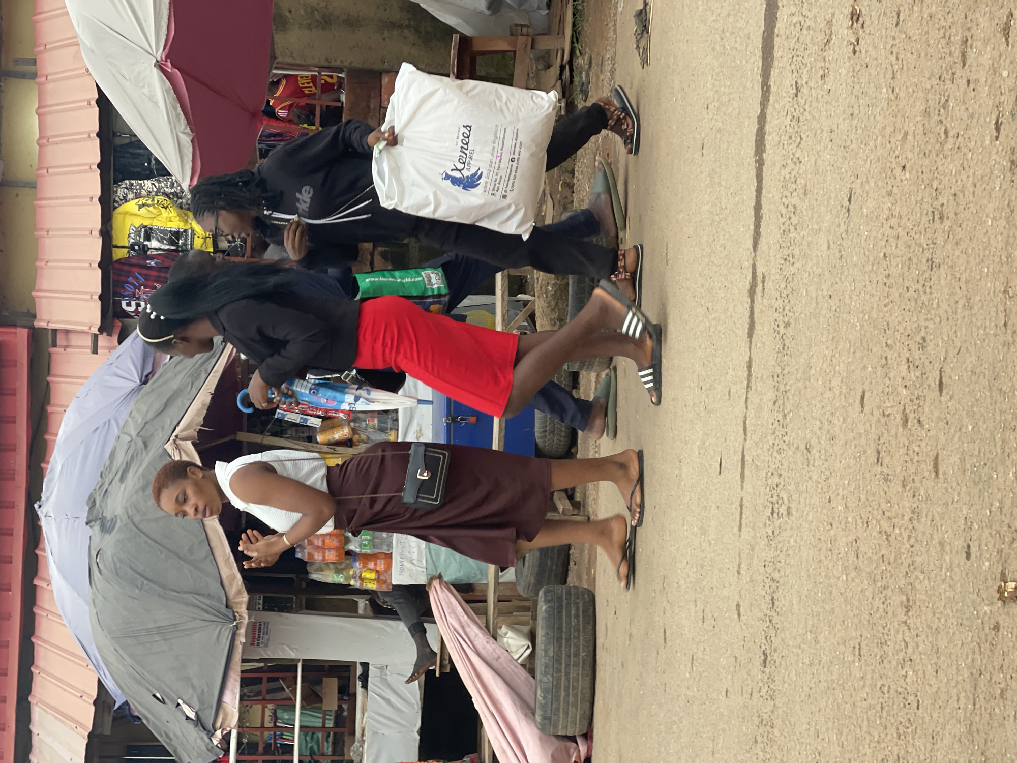 The image depicts a street scene in Abuja, Nigeria, likely a market area given the stalls and merchandise visible. In the foreground, three women are walking away from the camera. The woman on the left wears a white sleeveless top and a brown wrap skirt, carrying a black and gold purse. The middle woman wears a black jacket and a bright red skirt, with her hair in a ponytail adorned with a gold headband. The woman on the right wears a black hooded jacket and black pants, carrying a large white shopping bag with text and a blue bird logo.
Behind the women, market stalls are visible under large umbrellas. One stall displays rows of bottled drinks, and another shows clothing items, including a yellow jacket with black lettering and other garments. Tires are stacked on the ground near the stalls, and a person is seated in the background, partially obscured. The ground is paved with asphalt. The overall atmosphere suggests a bustling commercial environment.