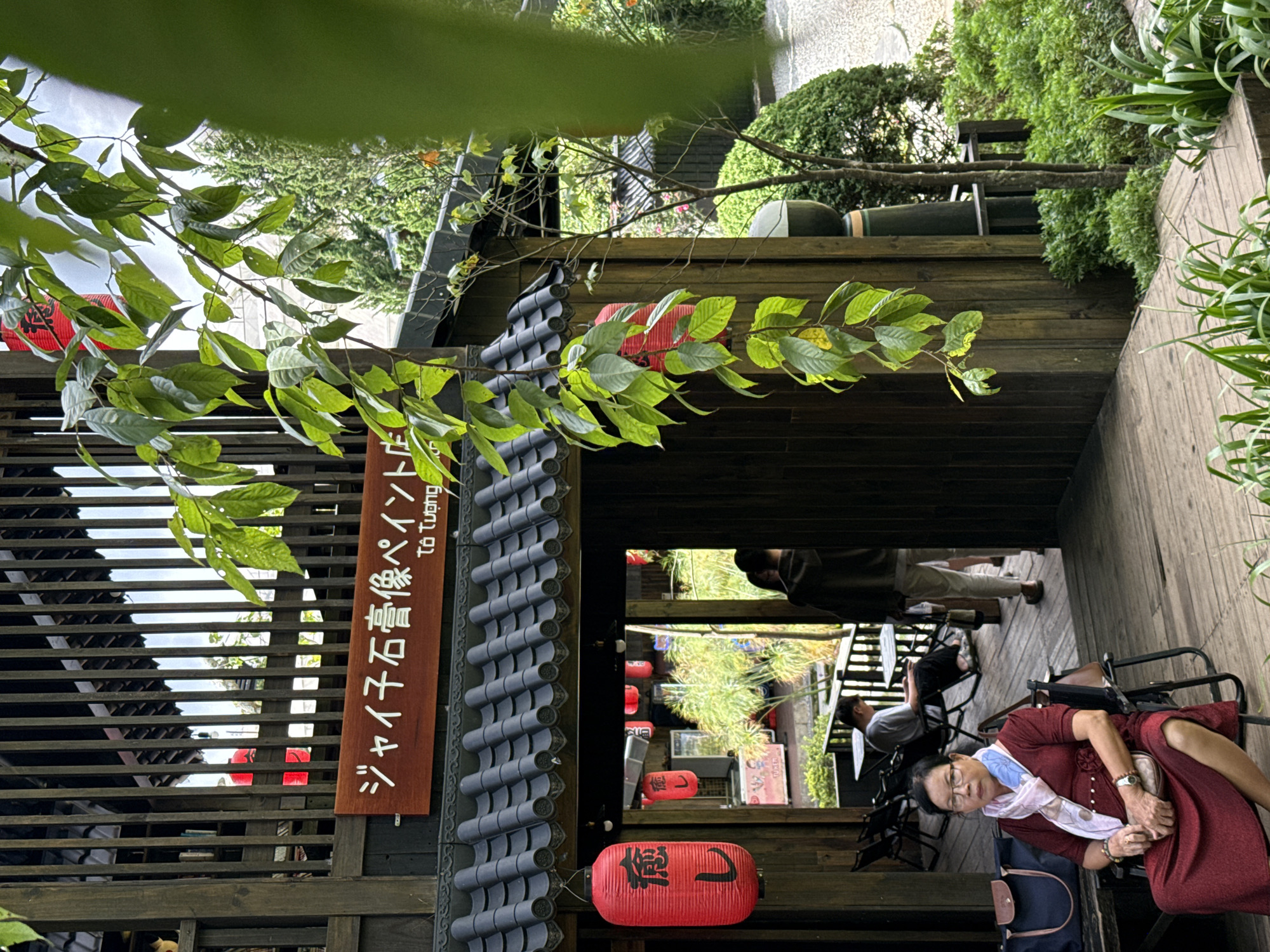 The image depicts a Japanese-themed structure in Cam Ly - Đà Lạt, Vietnam, with a prominent wooden building featuring a dark tiled roof. A horizontal wooden sign above the entrance displays Japanese characters "ジャイ子石膏像ペイント店" (Jaiko Sekkōzō Peinto-ten), translated below as "Tô Tượng" in Vietnamese, indicating a plaster statue painting shop.
In the foreground, an East Asian woman is seated on a dark chair on a wooden deck. She wears a long-sleeved, reddish-brown dress, clear-framed glasses, and a light blue and white patterned scarf. A wristwatch is on her left wrist, and a light brown handbag rests on her lap. To her left, a dark blue bag with brown straps is placed on a wooden bench.
To the left of the main entrance, a red cylindrical lantern hangs, bearing black Japanese characters "癒し" (iyashi - healing/relaxation). More red lanterns are visible inside the structure and through the vertical wooden slats of the upper story.
Further into the building, a male wearing a light grey t-shirt and dark pants is seated at a dark table with a dark chair. Another individual is partially visible entering the building from the right.
Lush green foliage surrounds the structure. A large branch with vibrant green leaves descends from the upper right, partially obscuring the view. Various green bushes and plants, including tall, thin blades of grass, are arranged around the wooden deck and along a light-colored path to the right. A small tree stands next to the building's right side. The sky is partially visible through the upper structure's slats.