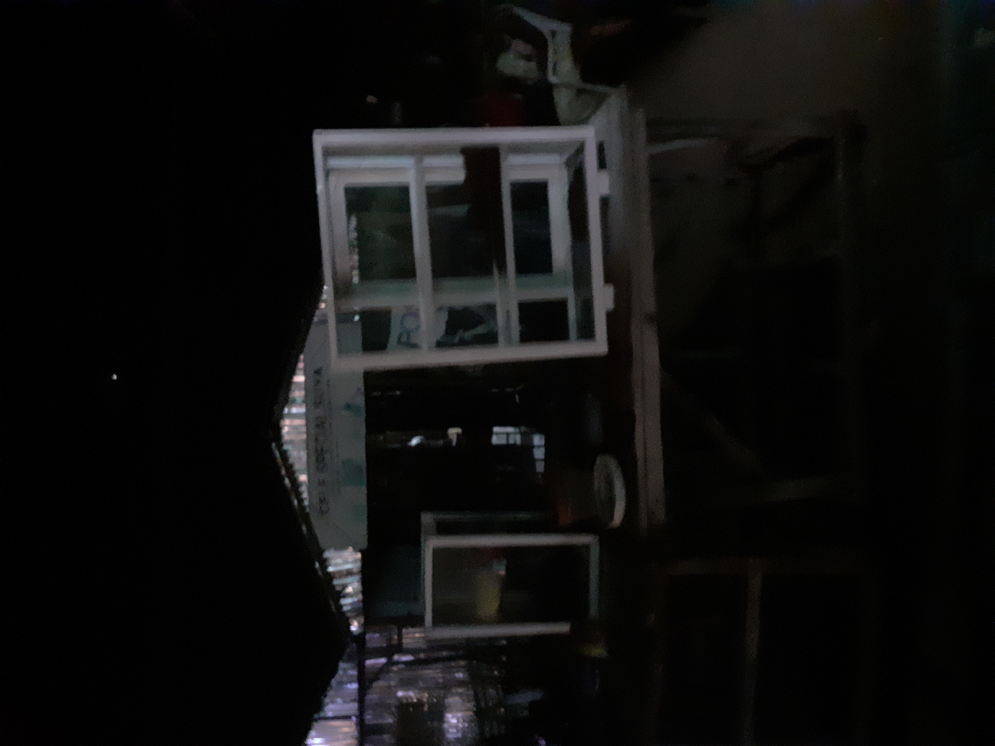 The image depicts a night scene in Jalingo, Nigeria. In the foreground, there are several objects placed on wooden surfaces, including two framed structures that appear to be windows or display cases. One is a tall, rectangular frame with multiple panes, and another is a similar frame placed horizontally on a surface. To the left, a shorter, vertically oriented framed object with a dark interior is visible. The background shows a partially covered structure, possibly a market stall or shop, with corrugated metal roofing. A sign with text, partially obscured, is visible on this structure. In the far right background, a person is seated on a chair, and another figure is partially visible. The overall lighting is dim, suggesting it is nighttime.