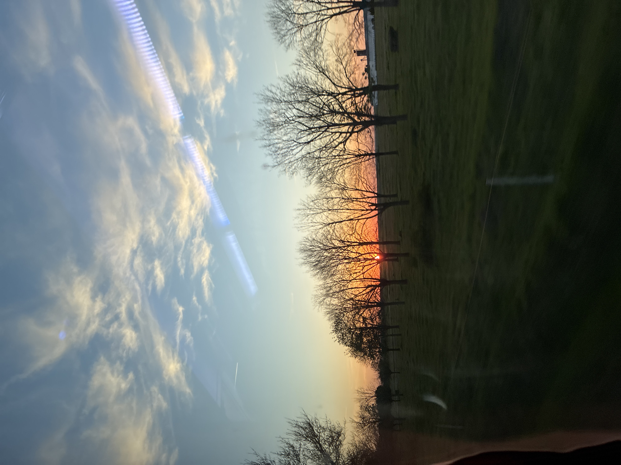 The image presents a serene rural landscape in Butan, Bulgaria, captured during either sunset or sunrise. The photograph appears to be taken from inside a vehicle, looking out through a window, as evidenced by distinct light streaks and reflections across the sky.
The sky transitions from a soft blue at the top to warm hues of orange and yellow closer to the horizon, where the sun is visible. Wispy white clouds are scattered across the blue sky, some catching the light of the sun. The sun itself is a vibrant orange-red orb, partially obscured by the intricate, bare branches of several deciduous trees. These trees are silhouetted against the bright sky, their leafless state suggesting late autumn, winter, or early spring. They are arranged in a linear fashion, possibly alongside a field or road.
Below the tree line, a grassy field stretches across the landscape, appearing green but somewhat subdued by the low light of the day. Faint structural elements, resembling fence posts or utility poles, are discernible within the field, particularly on the right side. The overall atmosphere conveyed is tranquil and picturesque. No text is visible within the media file.