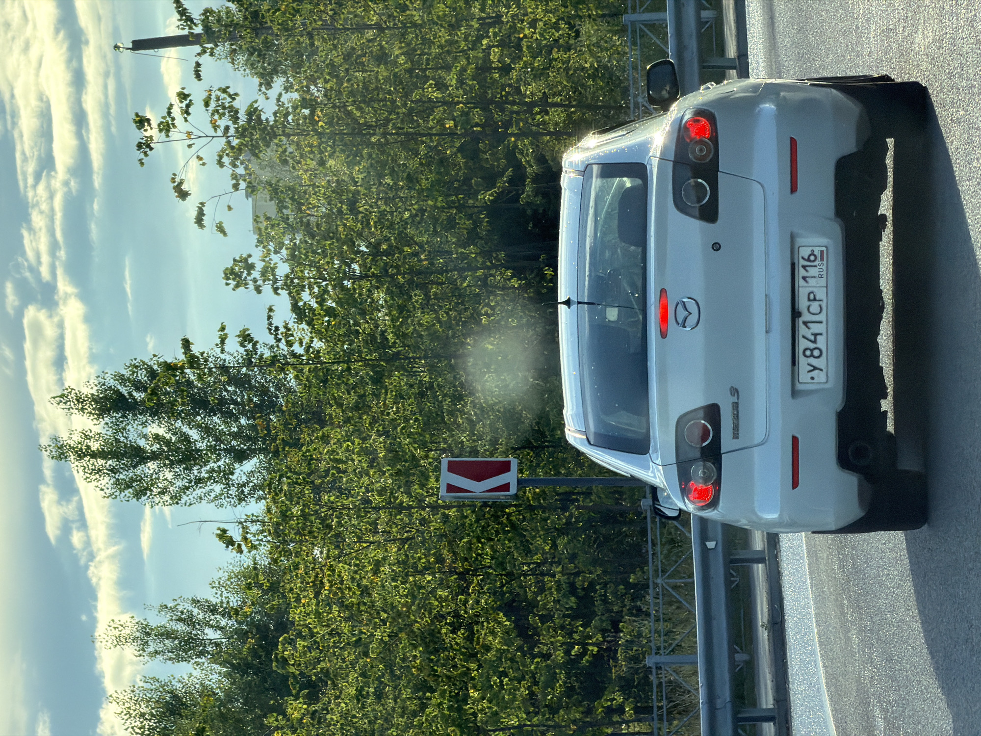 A rear view of a white 2010-2013 Mazda 3 sedan is captured from behind while driving on a paved road. The car is white with a red brake light illuminated. The license plate is visible and reads "y841cp 116". The car is surrounded by lush green trees and a bright blue sky with scattered clouds. A road sign with a red and white curved arrow is visible to the left of the car. The scene appears to be a highway or a scenic route. The lighting suggests it is either early morning or late afternoon. The car is positioned in the center of the frame, with the road stretching out in front of it. The overall impression is one of a peaceful drive through a natural landscape.