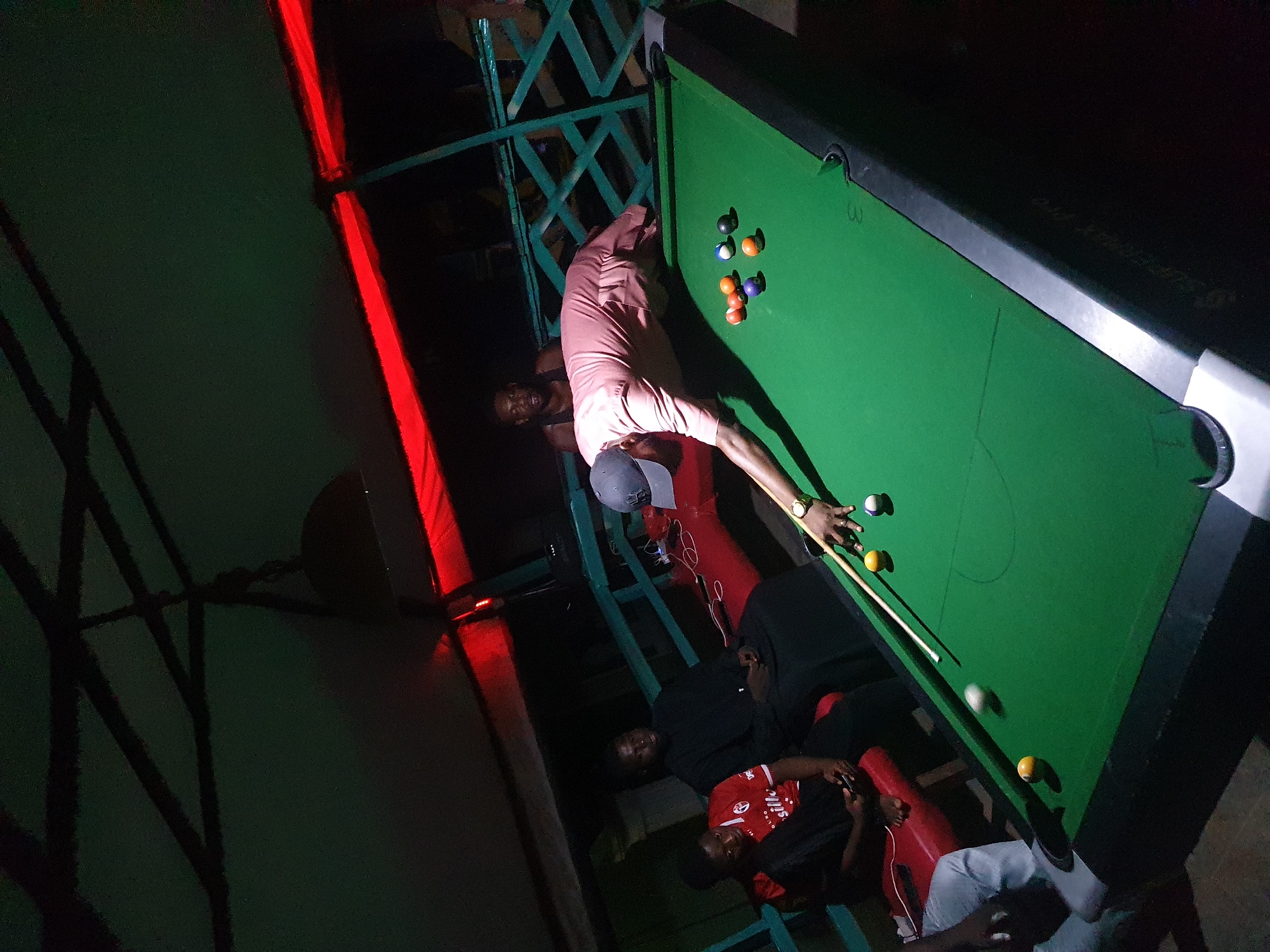 In a dimly lit interior, a group of men are playing pool on a green-felted table. One man, wearing a pink shirt and grey cap, is actively aiming the cue stick. Several other men are gathered around the table, some sitting on what appears to be a red bench-like structure. The lighting is a mixture of green and red hues, casting long shadows across the scene. A metal framework is visible near the pool table. Given the prompt's mention of Lokoja, Nigeria, it can be inferred that this scene is taking place in a pool hall in that city. The men's actions suggest a casual, social game of pool.