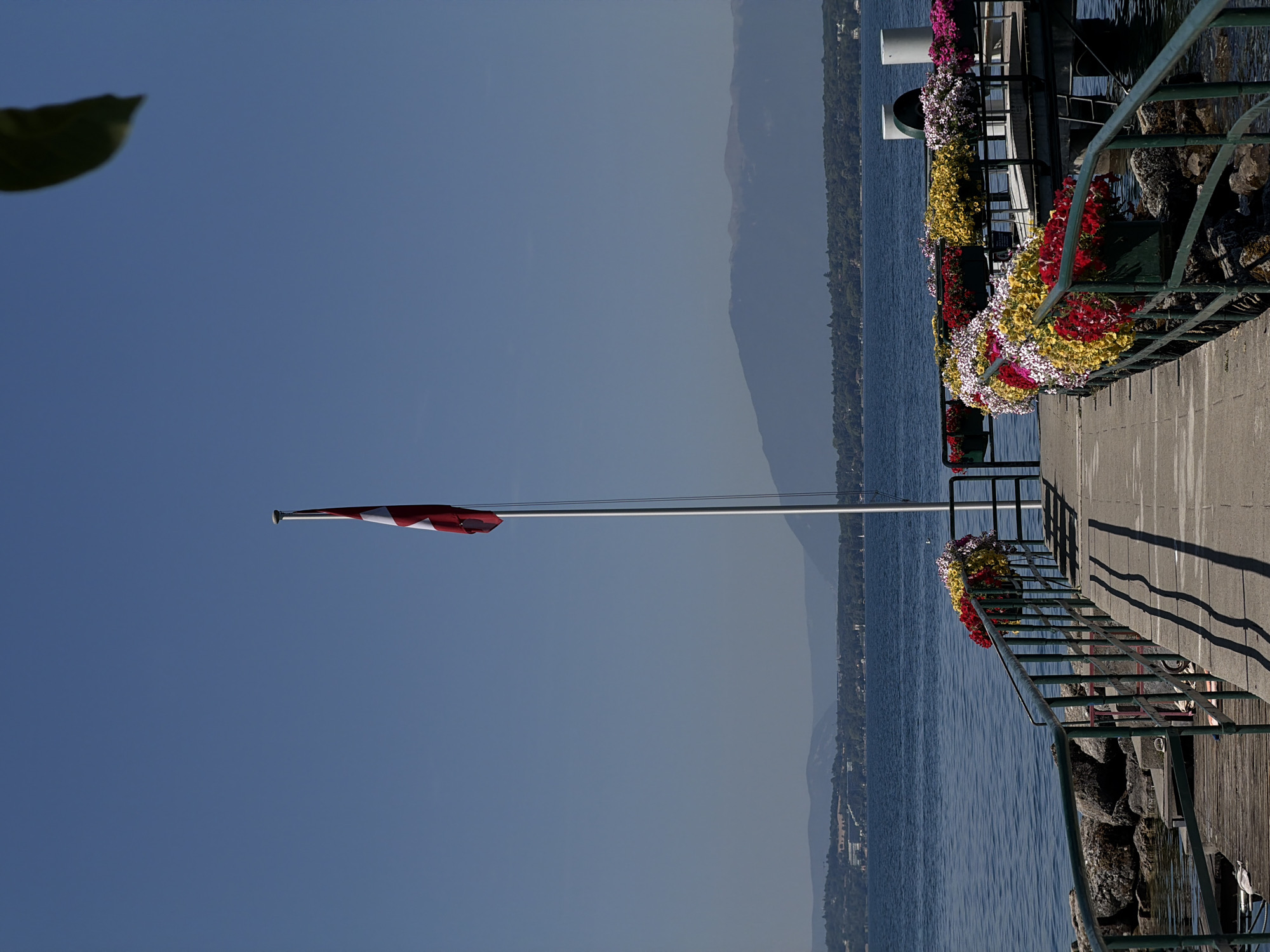 The image depicts a waterfront scene with a pier extending into a calm blue body of water, likely Lake Geneva, given the presence of mountains in the distance and the distinct architecture visible along the shoreline. A Swiss flag, red with a white cross, flies from a tall flagpole positioned on the pier. The pier itself is paved with light-colored material and features a green metal railing along its edge. Along the railing are several planters overflowing with a colorful array of flowers, including red, yellow, and white blooms.
To the left of the pier, a rocky shoreline is visible with weathered wooden structures. The water is clear, and the sky is a bright, cloudless blue. In the background, a distant shoreline is lined with trees and buildings, with rolling hills and mountains forming a natural backdrop. The overall impression is one of a serene and well-maintained public space, likely a popular spot for leisure and viewing the lake and surrounding landscape.