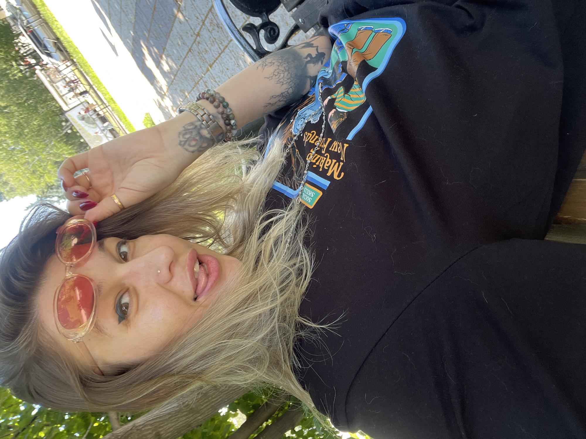 A woman with blonde hair, wearing a black t-shirt with a graphic print and red-tinted sunglasses, is taking a selfie outdoors. She is sitting on a bench, with trees and a sunny outdoor seating area visible in the background. She has tattoos on her arm and is wearing rings and bracelets. Her tongue is sticking out of her mouth, and she is touching her sunglasses with her right hand. The image is taken from a high angle, looking down at the woman.