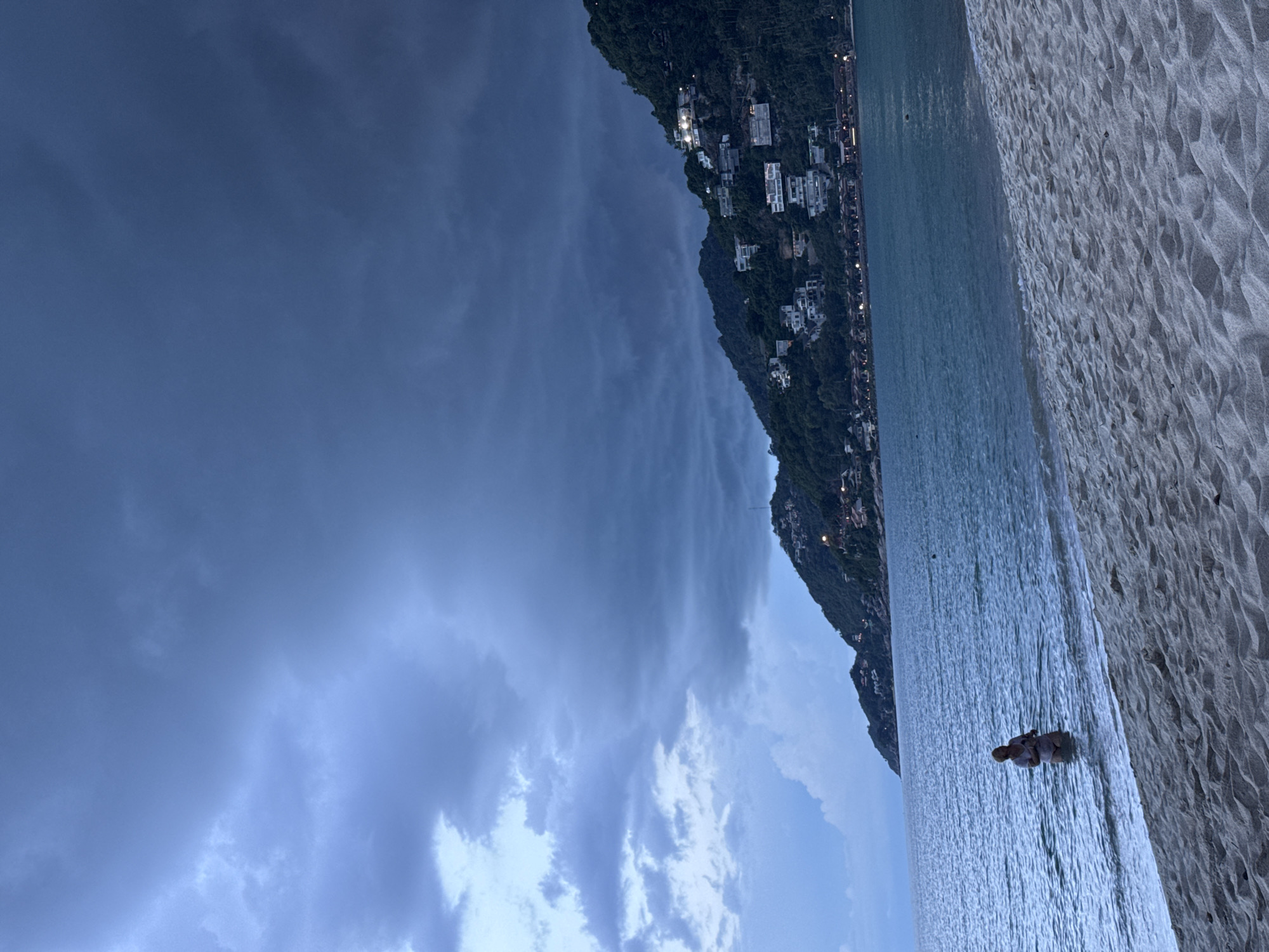 The image depicts a serene beach scene at Baan Chaweng Noi, Thailand. The sky is overcast with dramatic, dark clouds, suggesting either twilight or an impending storm. The sea is calm and a muted blue, gently lapping the shore. A strip of light-colored sand forms the foreground, showing the texture of the grains.
On the left side of the image, a steep, tree-covered hillside rises, dotted with numerous white buildings that appear to be houses or villas, many illuminated by lights. This suggests a densely populated residential or resort area built into the landscape. The lights along the coastline indicate it's either evening or a very overcast day.
In the water, near the shore, a single person is visible, partially submerged. They appear to be sitting or kneeling, facing away from the camera, seemingly enjoying the water despite the moody weather. The overall mood is tranquil and somewhat dramatic due to the contrast between the dark sky and the illuminated buildings. There is no visible text in the image.