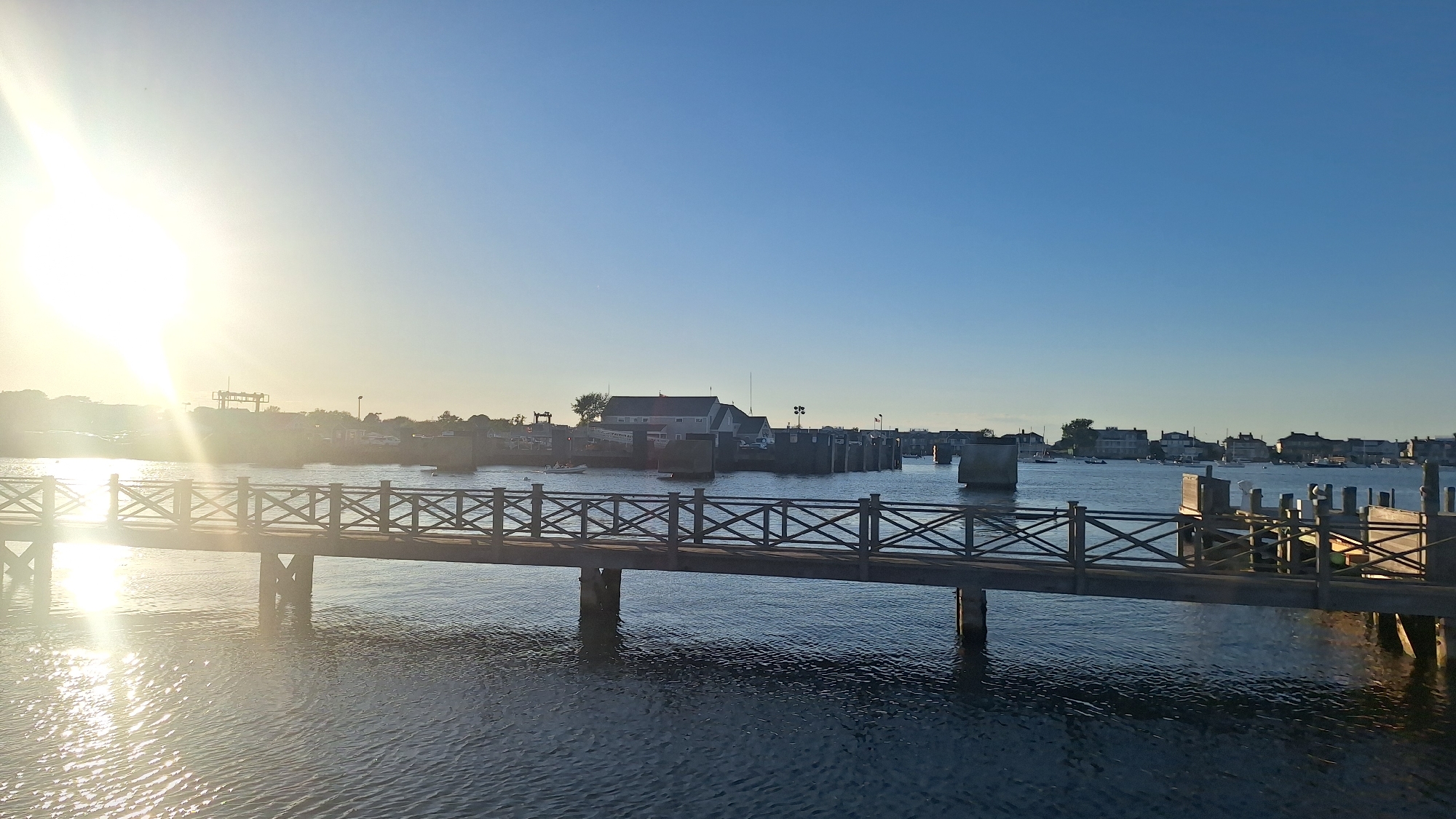 The image shows a wooden pier extending across a body of water. The pier has a decorative railing with crisscrossed wooden beams. The water is dark blue and reflects the sunlight. In the background, there are buildings and boats, suggesting a harbor or waterfront area. The sky is a clear, bright blue. The sun is visible on the left side of the image, casting a strong glare. The overall scene depicts a peaceful waterfront setting.