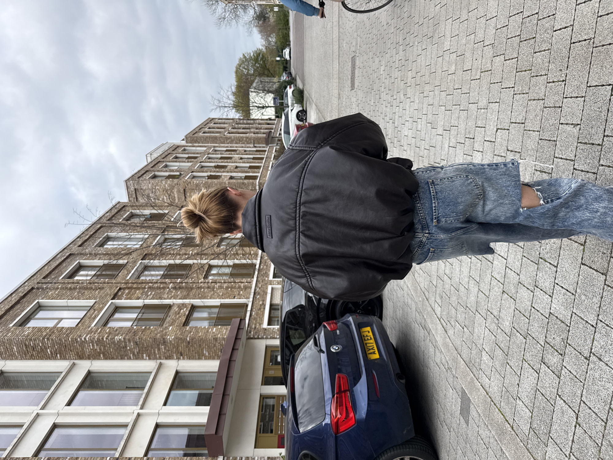 A person stands beside a dark-colored car parked on a paved street next to a multi-story brick building in London, United Kingdom. The person is wearing a dark jacket and ripped jeans, their hair in a bun. The car has a yellow license plate with the registration "AX17 EFJ". Other cars are parked further down the street, and a person on a bicycle is visible in the distance. The sky is overcast. The building features multiple rows of windows with a pattern of brick and white trim. The paving stones on the street are rectangular and arranged in a herringbone pattern.