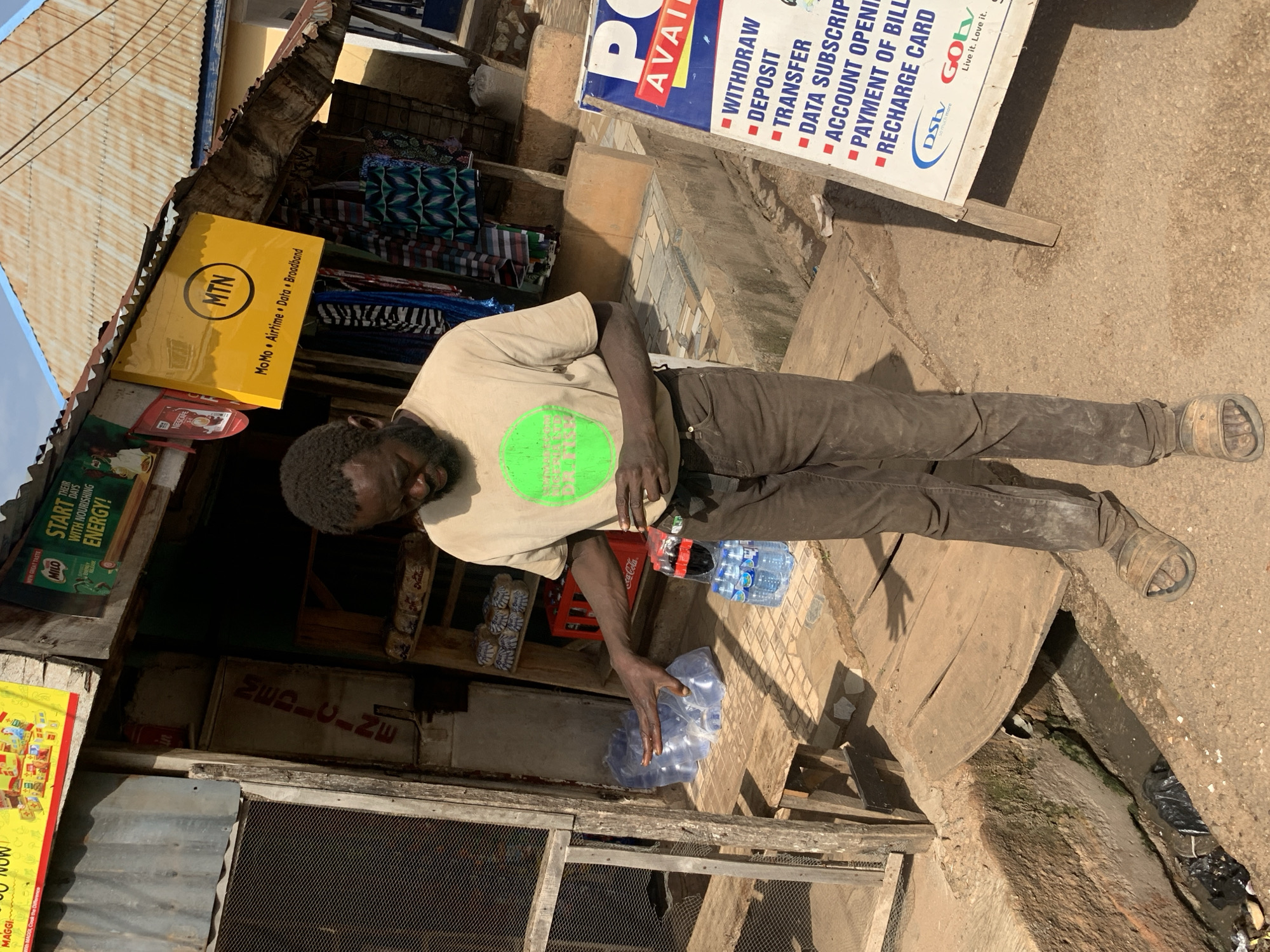 In Gura Top, Nigeria, a dark-skinned man with a beard stands outdoors in front of a small shop. He is wearing a light-colored t-shirt with a green circular graphic and dark brown pants. His left hand is outstretched, pointing towards several large plastic bottles of water and a six-pack of bottled drinks. Behind him, a wooden shelf holds loaves of bread. Above the shop entrance, a yellow sign displays the MTN logo and services like "MoMo • Airtime • Data • Broadband". A red Coca-Cola crate is visible behind the man. To the right of the scene, a blue and white sign lists financial services such as "WITHDRAW", "DEPOSIT", "TRANSFER", "DATA SUBSCRIPTION", "ACCOUNT OPENING", "PAYMENT OF BILLS", and "RECHARGE CARD", along with the DStv and GOtv logos. The environment appears to be a street scene with a drainage ditch to the left of the man.