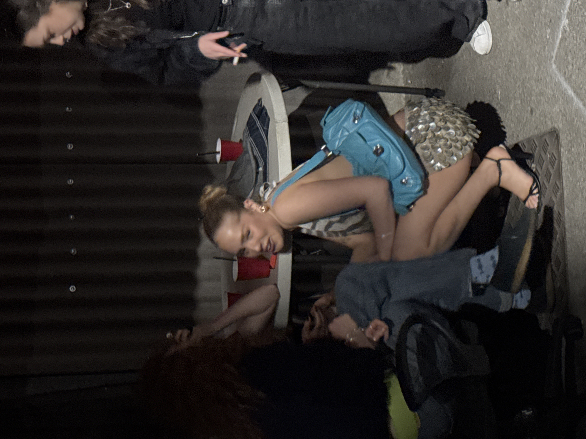 The image shows a group of individuals in an outdoor setting, possibly a gathering or event, with an urban environment suggested by the surrounding structures. A woman is positioned near a white folding table with red cups and a blue purse. She is wearing a dress with a sequin skirt and strappy sandals. Several other individuals are present, some interacting with the table and others obscured by shadow. The setting appears to be London, United Kingdom.