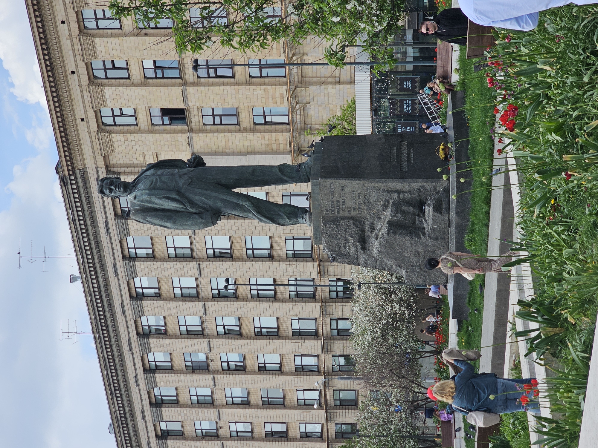 The image shows a full-length statue of Vladimir Mayakovsky in Moscow, Russia, set against a tall, brick building. The statue, composed of a dark material, stands atop a large stone pedestal. A sidewalk runs in front of the statue, bordered by flowerbeds with red flowers. People are seen on benches and walking in the background, and a ladder is visible near a building's glass facade. The environment includes foliage, trees, and a partly cloudy sky.