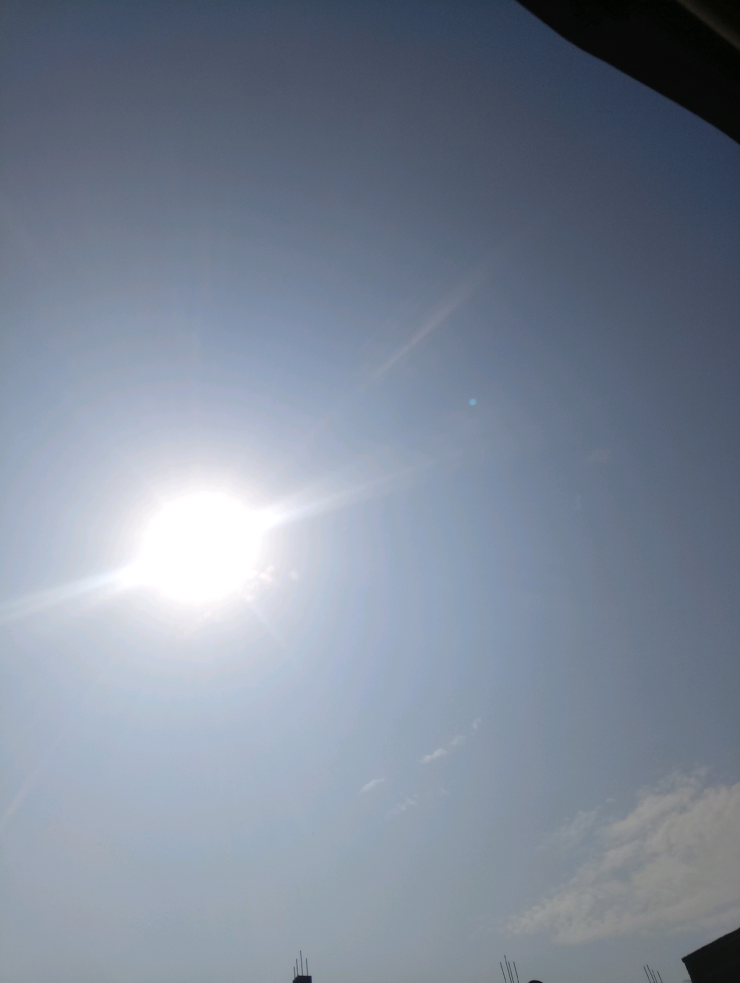 The image captures a bright, sunny sky over Chahania, India. The sun is prominently visible, a dazzling orb with rays of light extending outwards, casting a bright glow. The sky is a light blue, with a few wispy white clouds scattered across it. In the lower portion of the image, the dark silhouettes of building structures and what appear to be utility poles are visible against the bright sky. The overall impression is one of a clear, warm day.