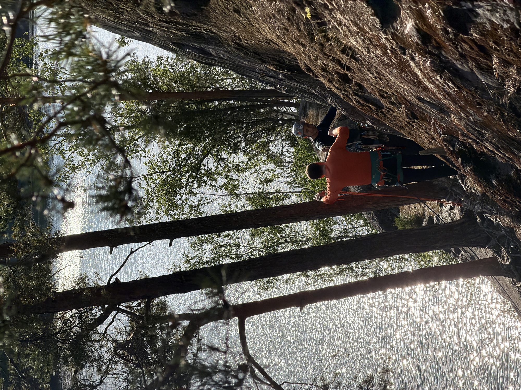 Two individuals are rock climbing on a steep, rocky incline near a body of water, likely a lake or river, in Ruch'i, Russia. The person in the foreground, wearing an orange long-sleeved shirt and a climbing harness, is facing away from the camera and appears to be preparing to climb. The second individual, wearing a helmet and a dark long-sleeved shirt, is positioned slightly behind and to the right, also equipped with a climbing harness. Both individuals are attached to climbing ropes. The rocky surface is textured and appears to be covered in moss and small vegetation in places. The background features a sun-dappled body of water with sparkling reflections, framed by the trunks of tall trees and green foliage. The overall lighting suggests a bright, sunny day.